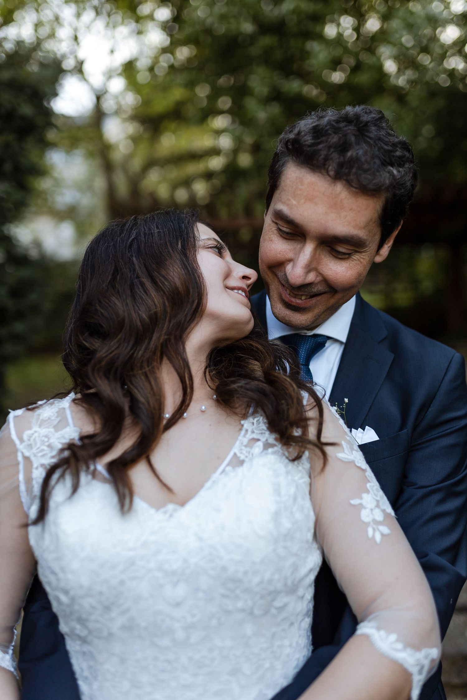 A bride in a white lace dress and groom in navy suit share intimate moments during an outdoor wedding photoshoot.