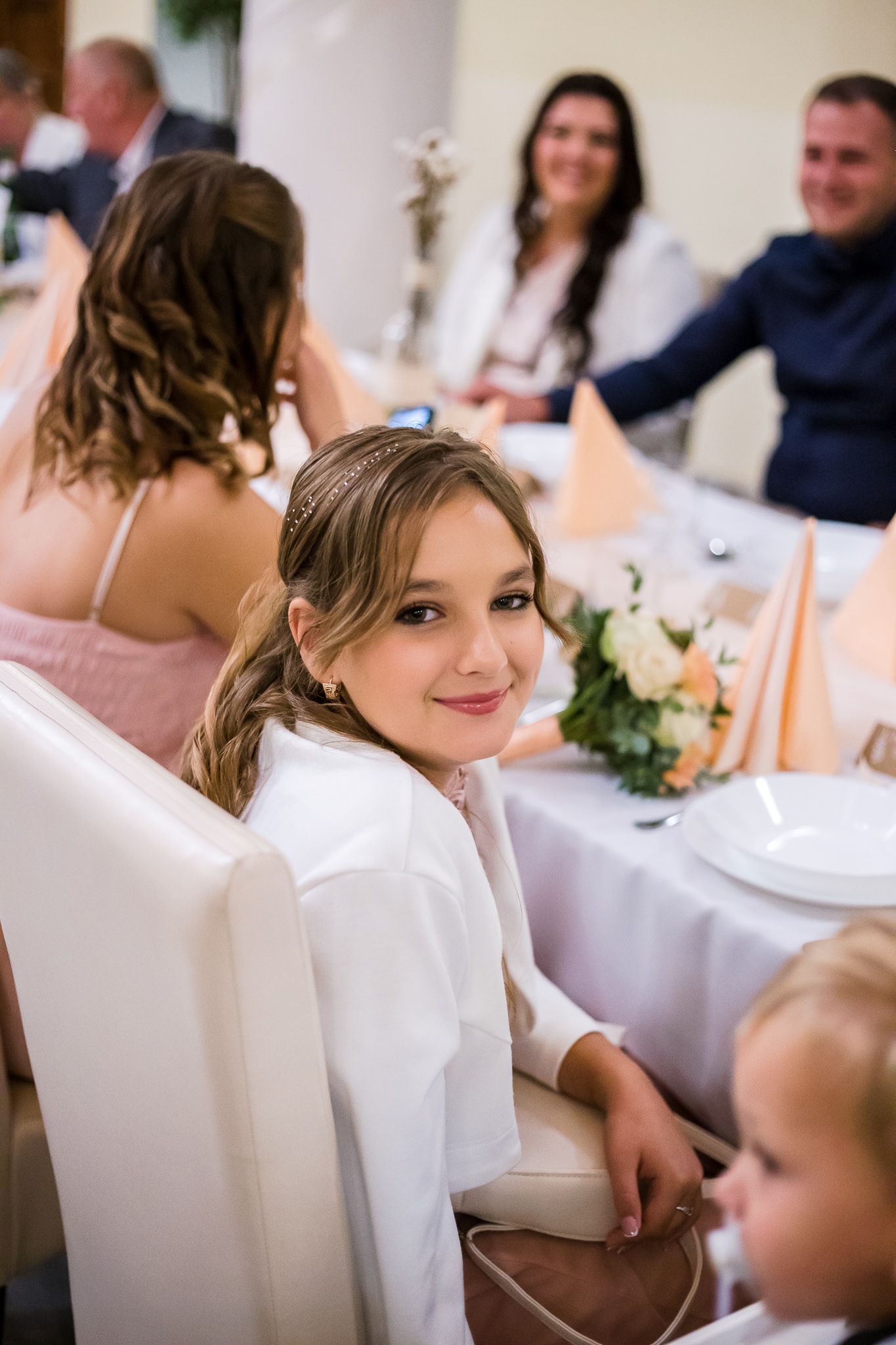 A guest in a white top sits at an elegant table during a formal dining event with other attendees in the background.