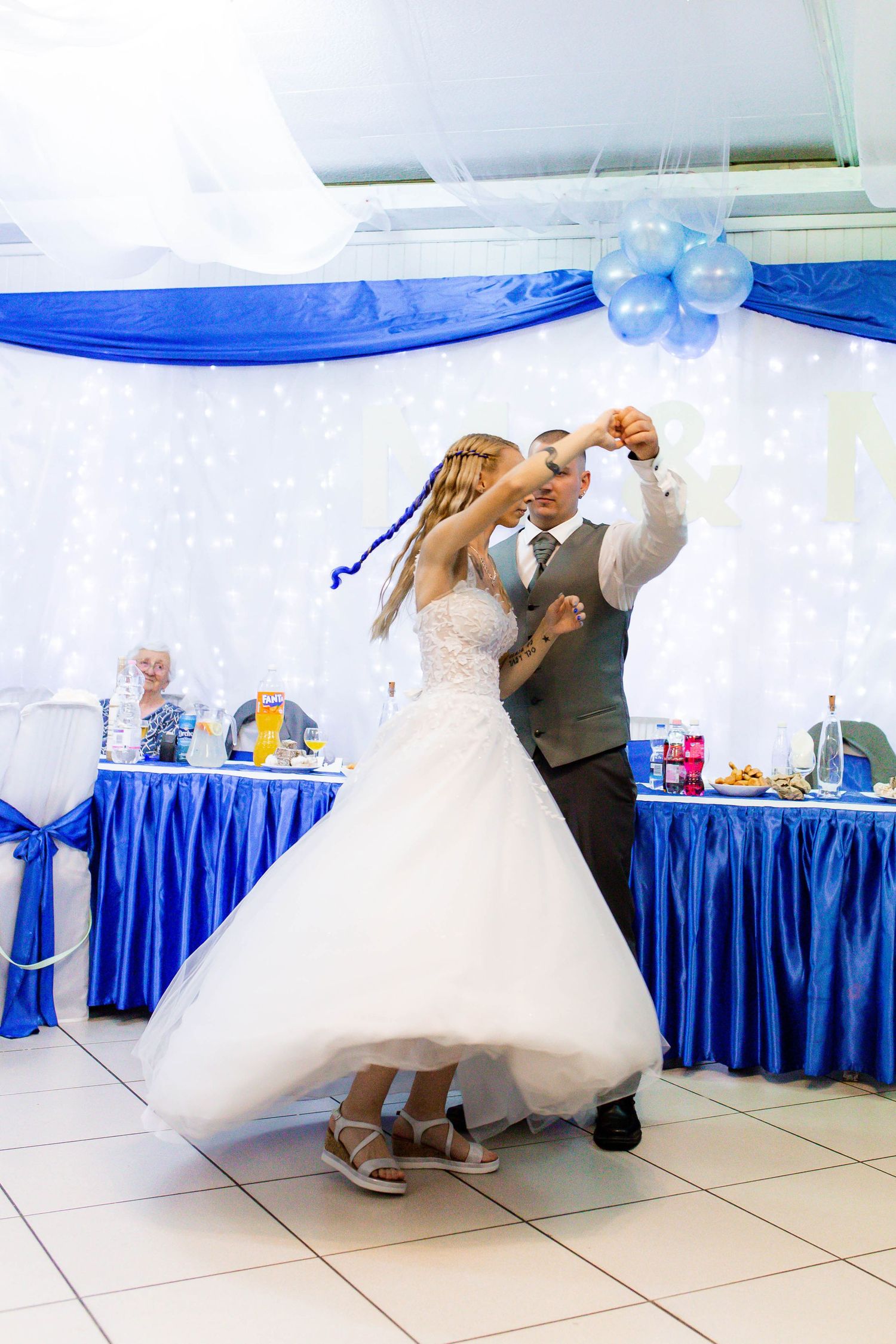 A couple performs a romantic dip dance move in formal wedding attire on a white dance floor with blue decorations.