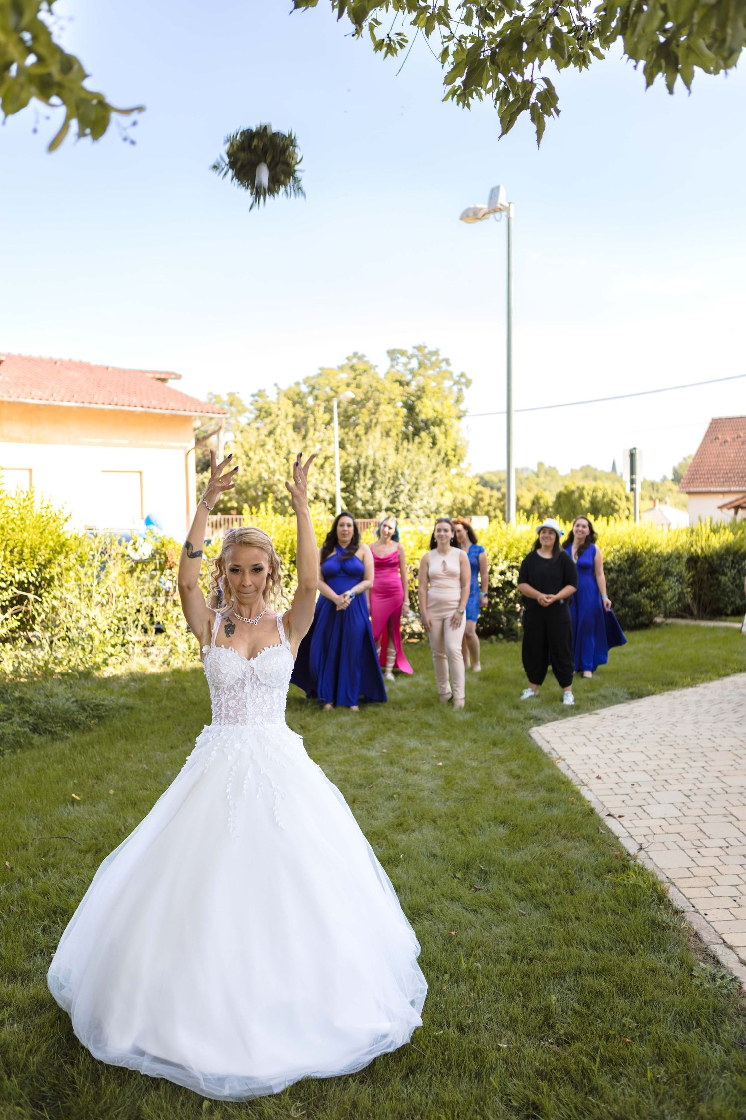A bride in a white ball gown throws her bouquet during an outdoor wedding ceremony while guests watch in the background.