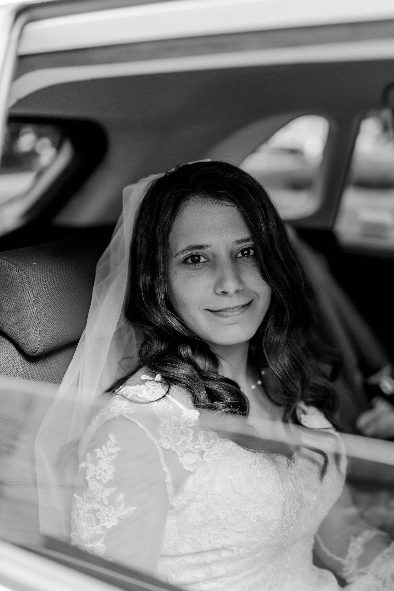 A bride in a white lace wedding dress sits elegantly in a car on her special day.