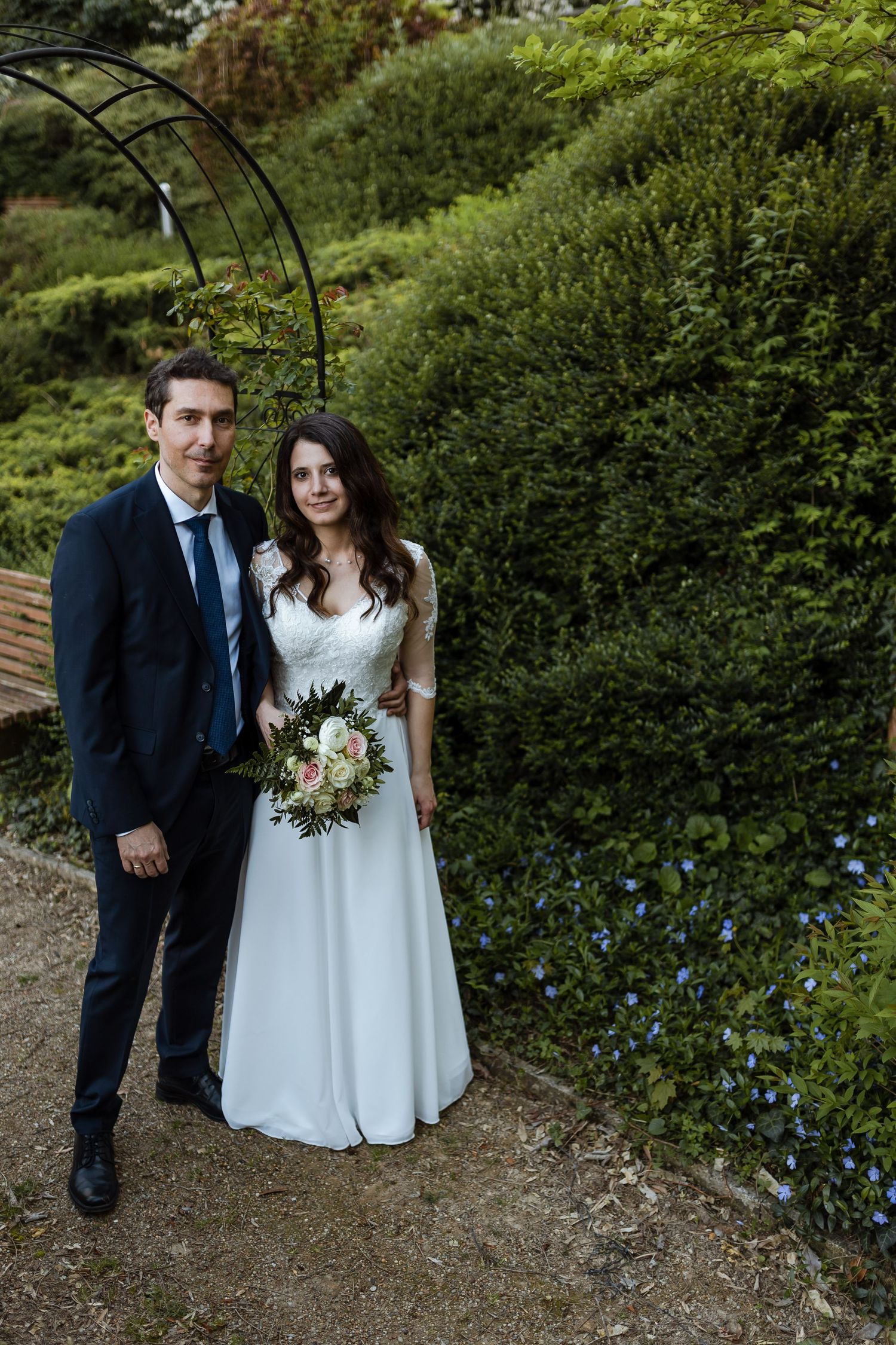 A couple in wedding attire poses in a garden setting with lush greenery and a metal arch structure.