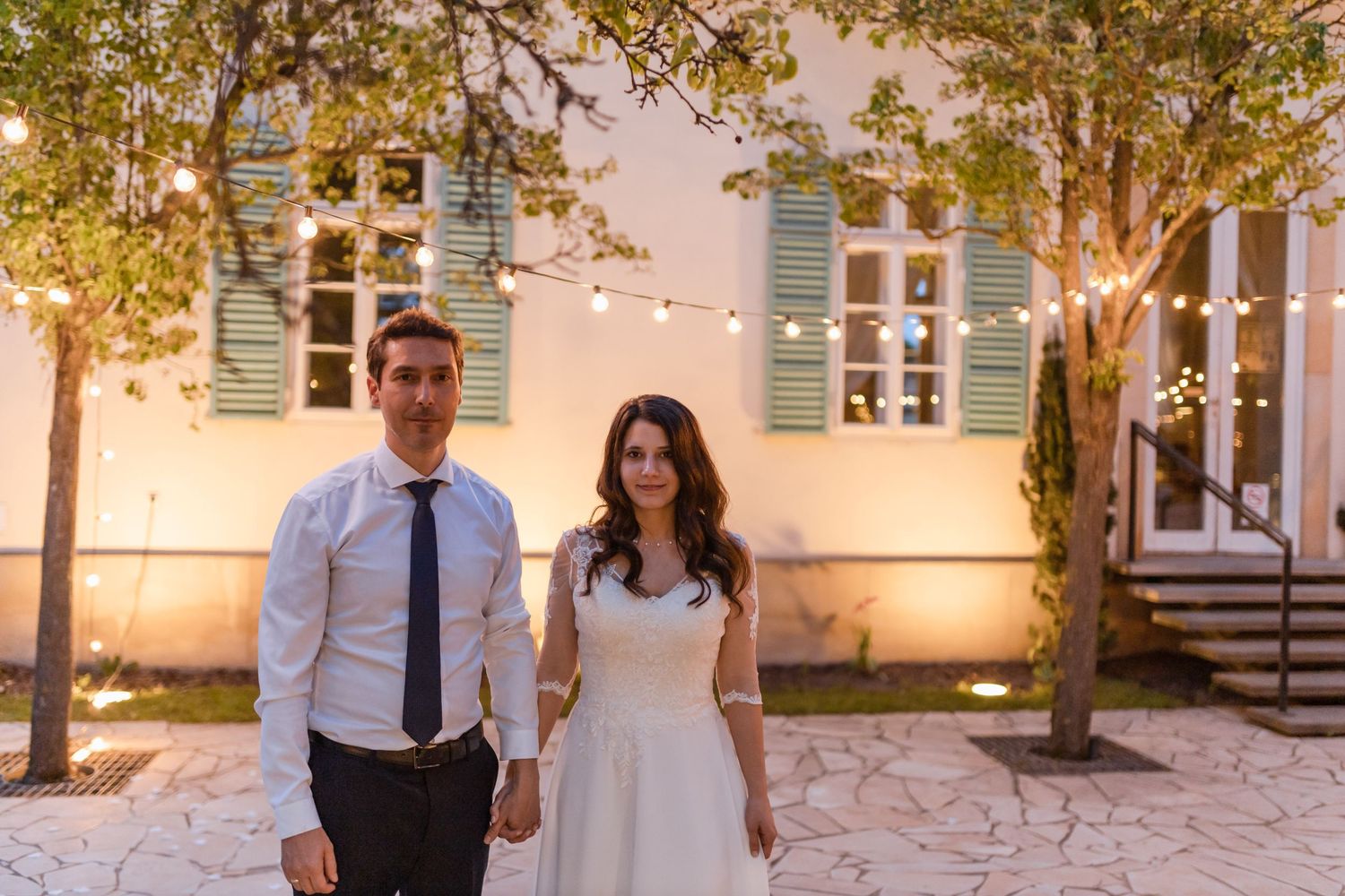 A couple stands together in a romantic outdoor courtyard setting with white stone walls and green shutters.