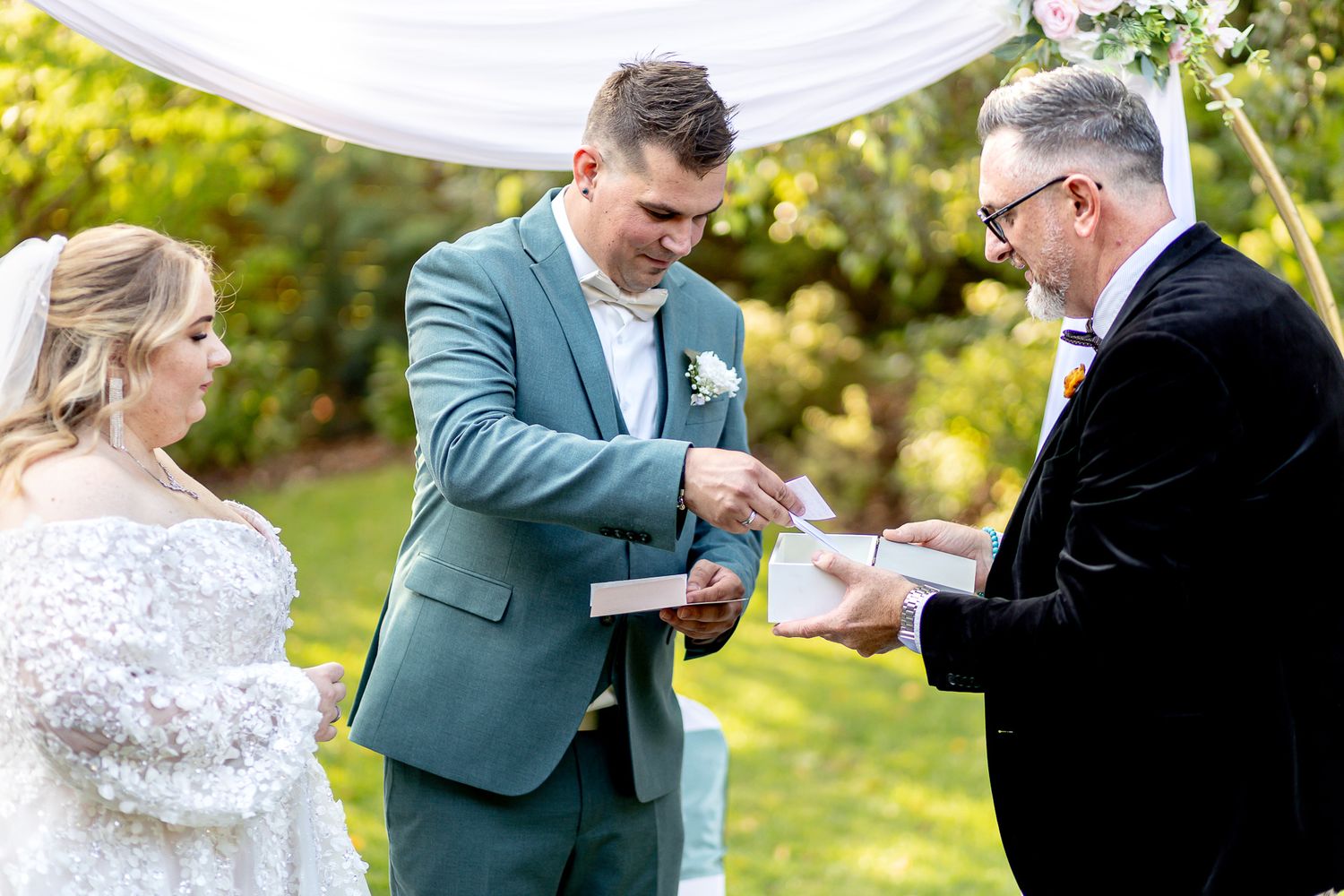 A couple participates in a wedding ceremony outdoors under a white canopy on a sunny day.