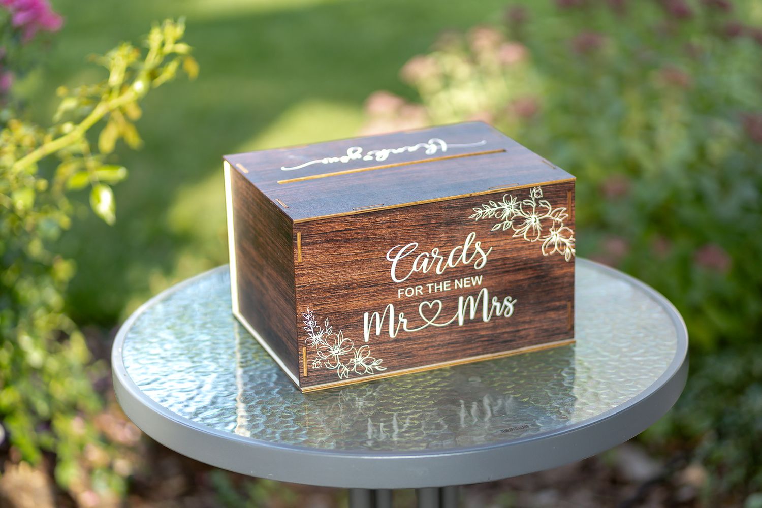 A rustic wooden wedding card box with 'Mr & Mrs' text sits on a silver table in a garden.