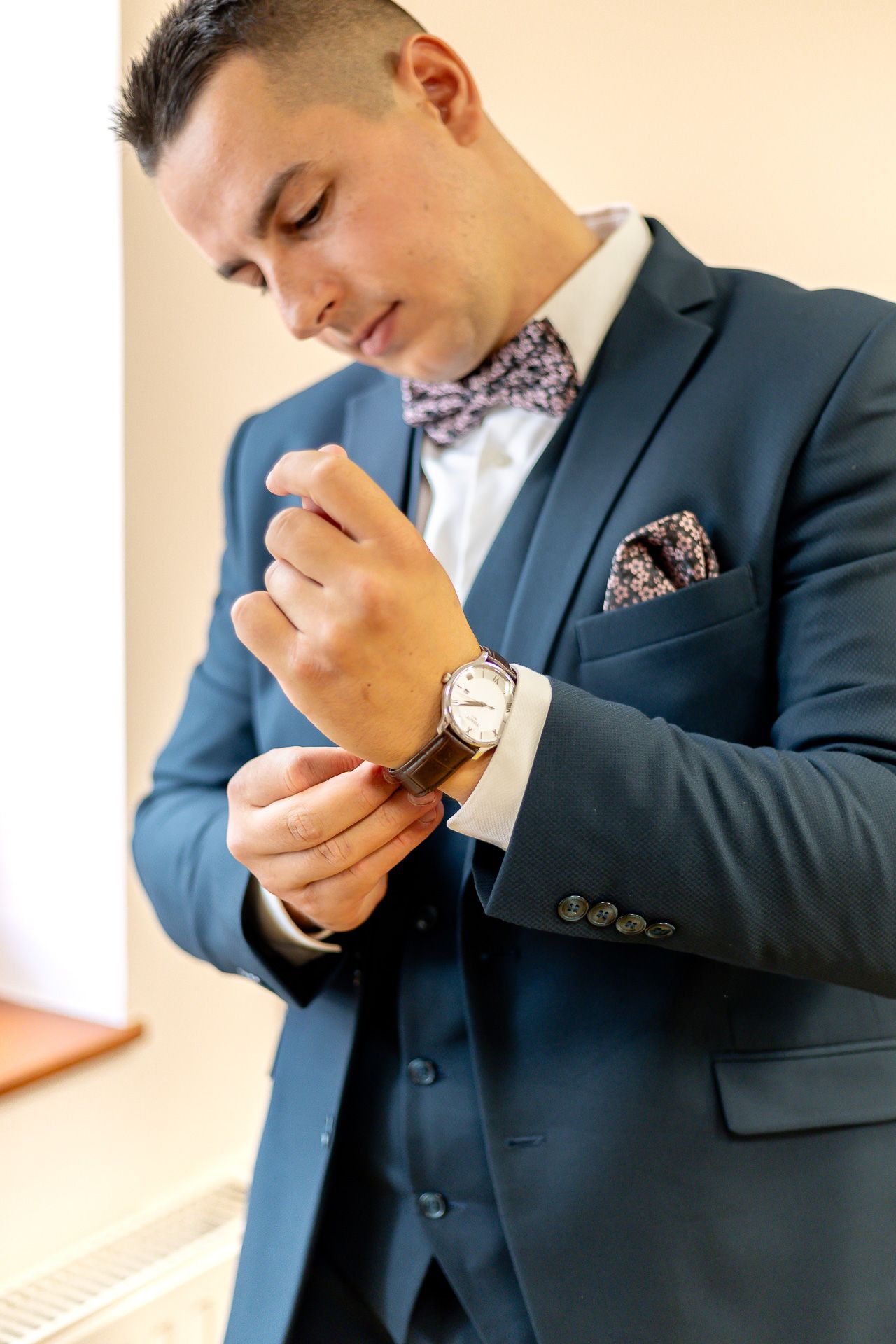 Person in navy suit adjusts watch on wrist while wearing floral pocket square and formal attire.