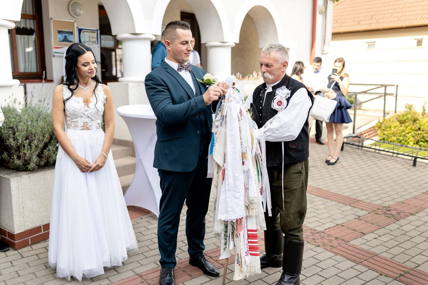 A group of wedding guests and bridal party members gather outside a church building with white arches.