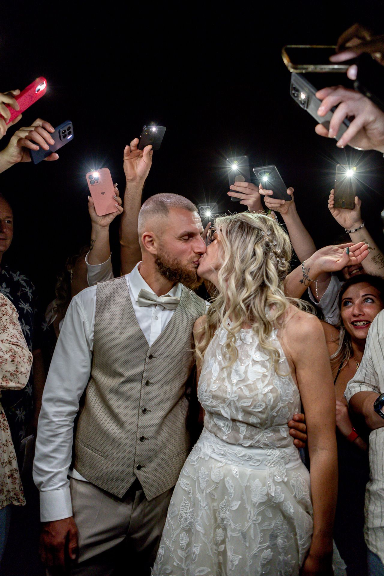 Wedding couple dances surrounded by guests holding up cell phones with flashlights in a dark reception venue.