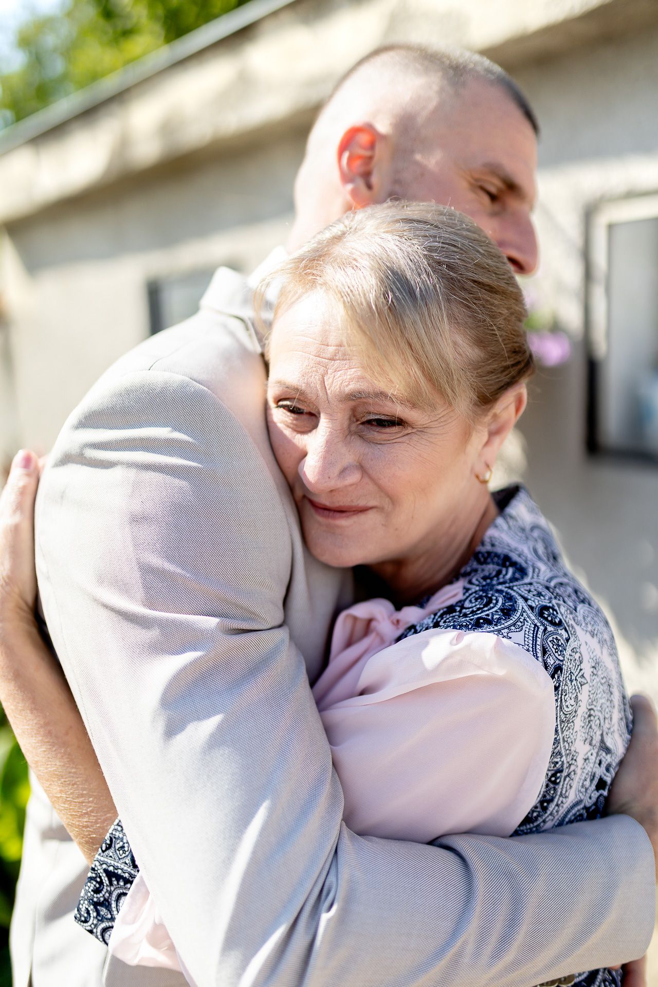Two people share a tender embrace outdoors on a sunny day, with one wearing a patterned scarf and pink top.