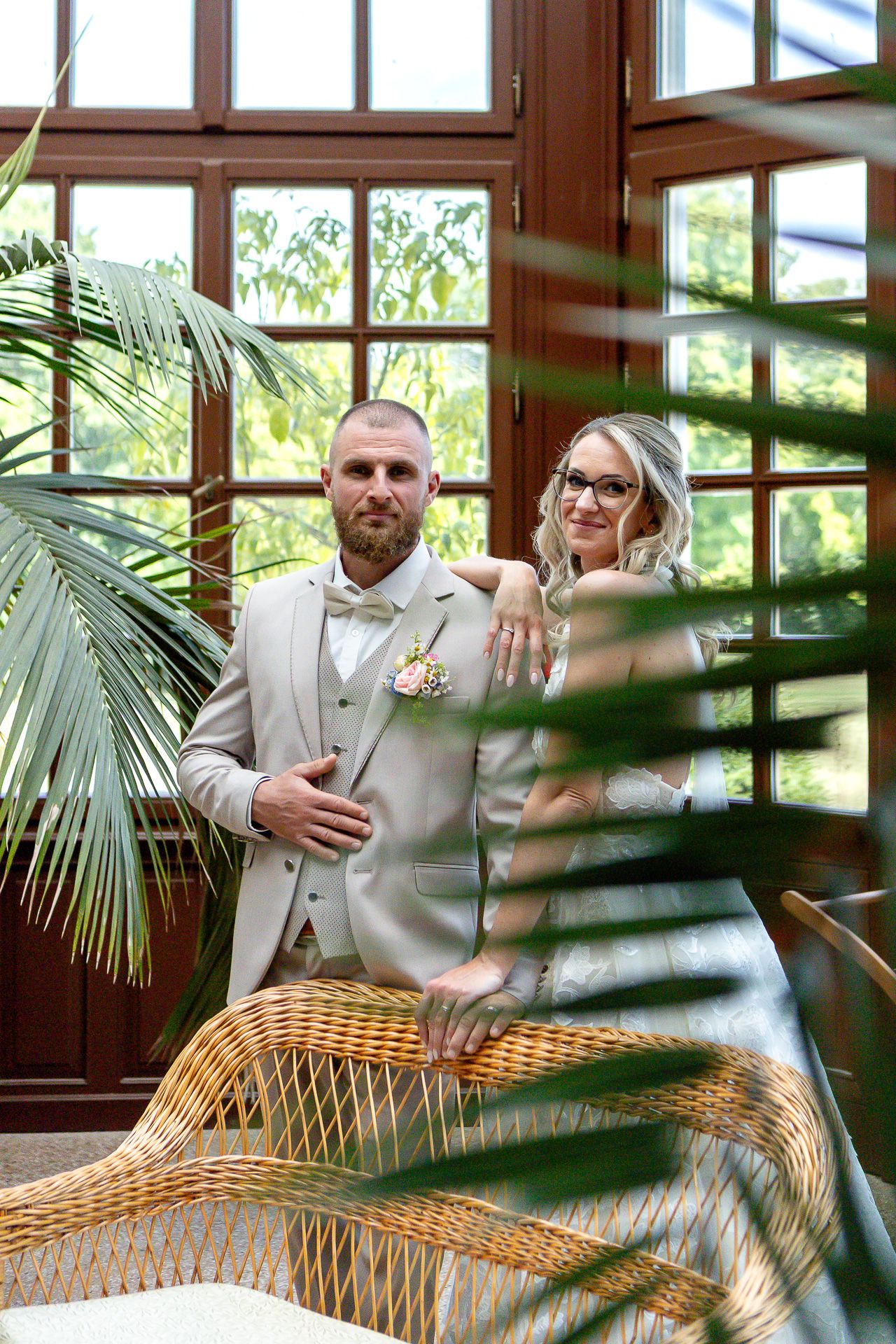 A couple poses together in a tropical conservatory setting with palm fronds and wicker furniture.