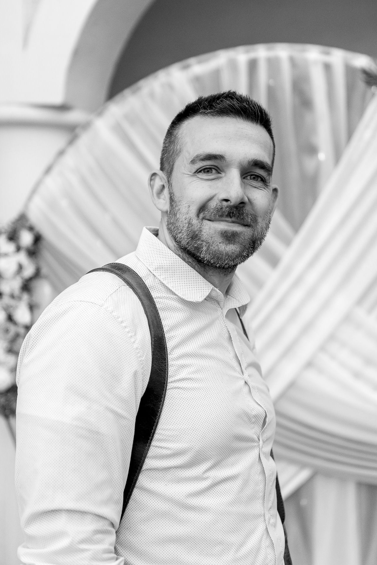 A person wearing a white collared shirt and black suspenders smiles while looking to the side in a bright setting.