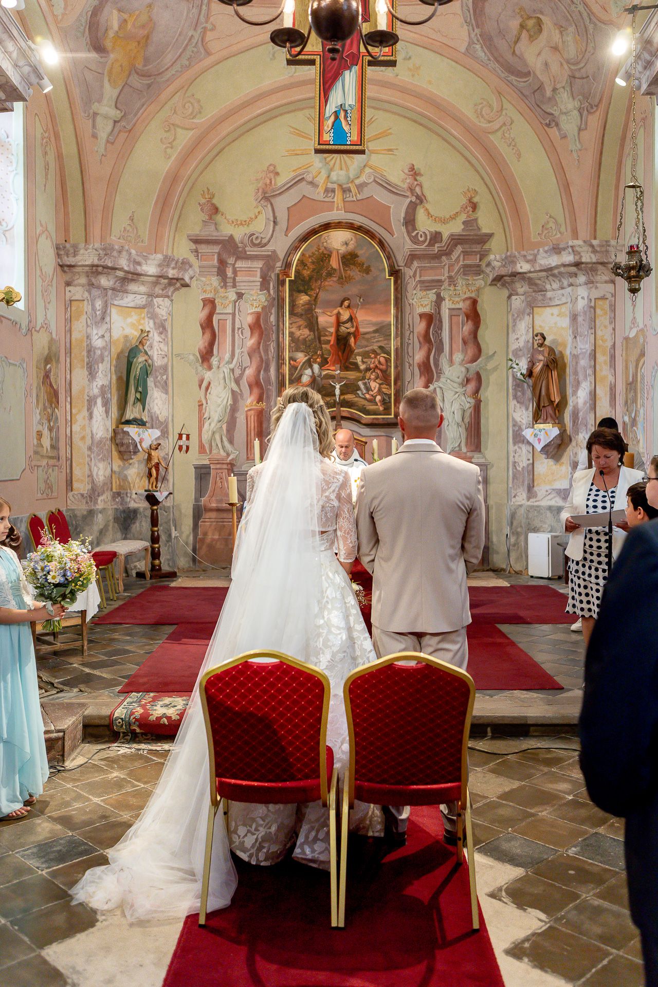 A wedding ceremony takes place inside an ornate church with pink frescoed walls and red velvet chairs.