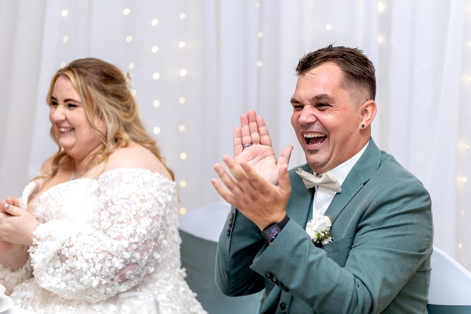 A bride and groom share a joyful moment during their first dance at a wedding reception with twinkling lights behind them.