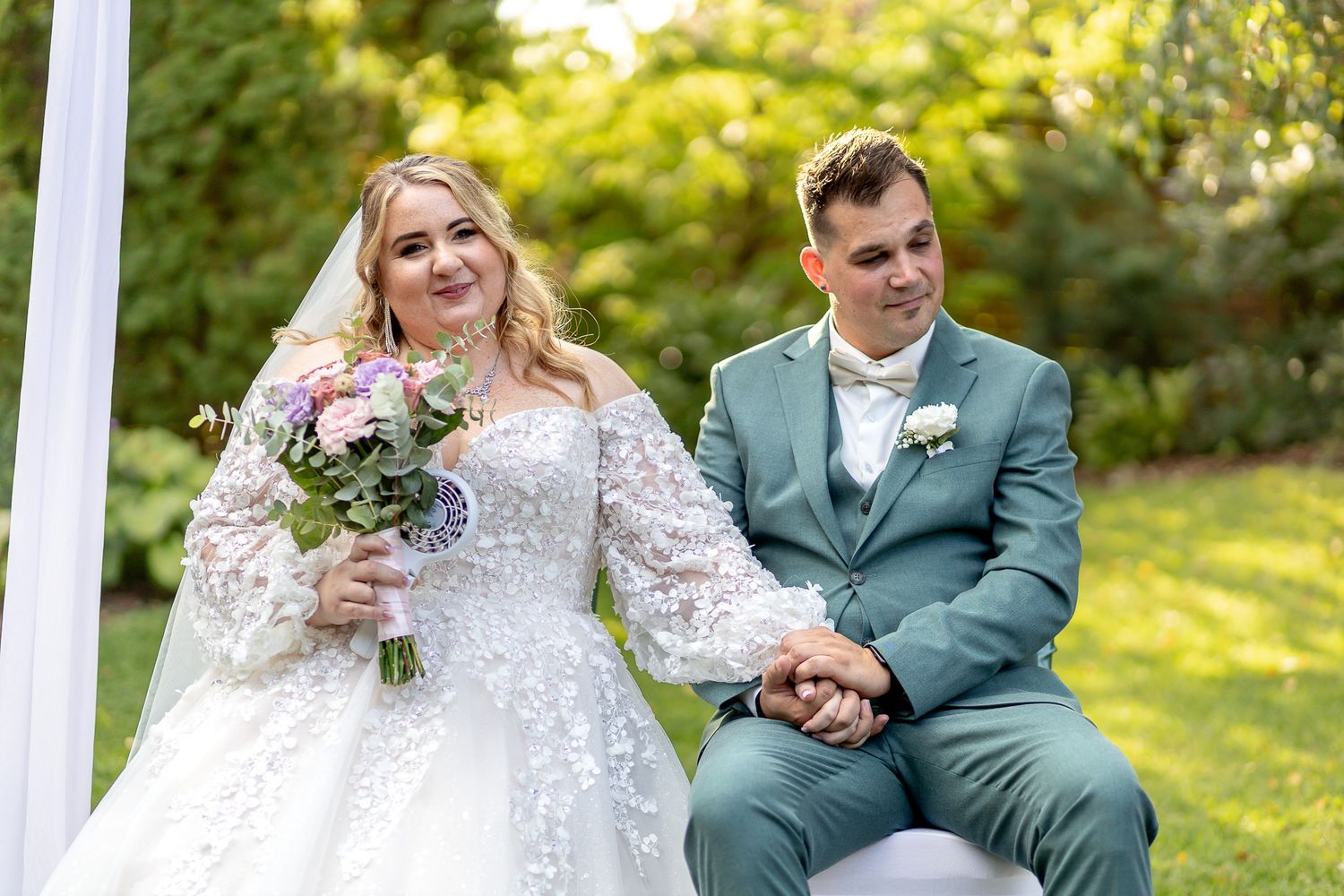 Bride and groom sit together on white porch swing in garden setting during summer wedding photoshoot.