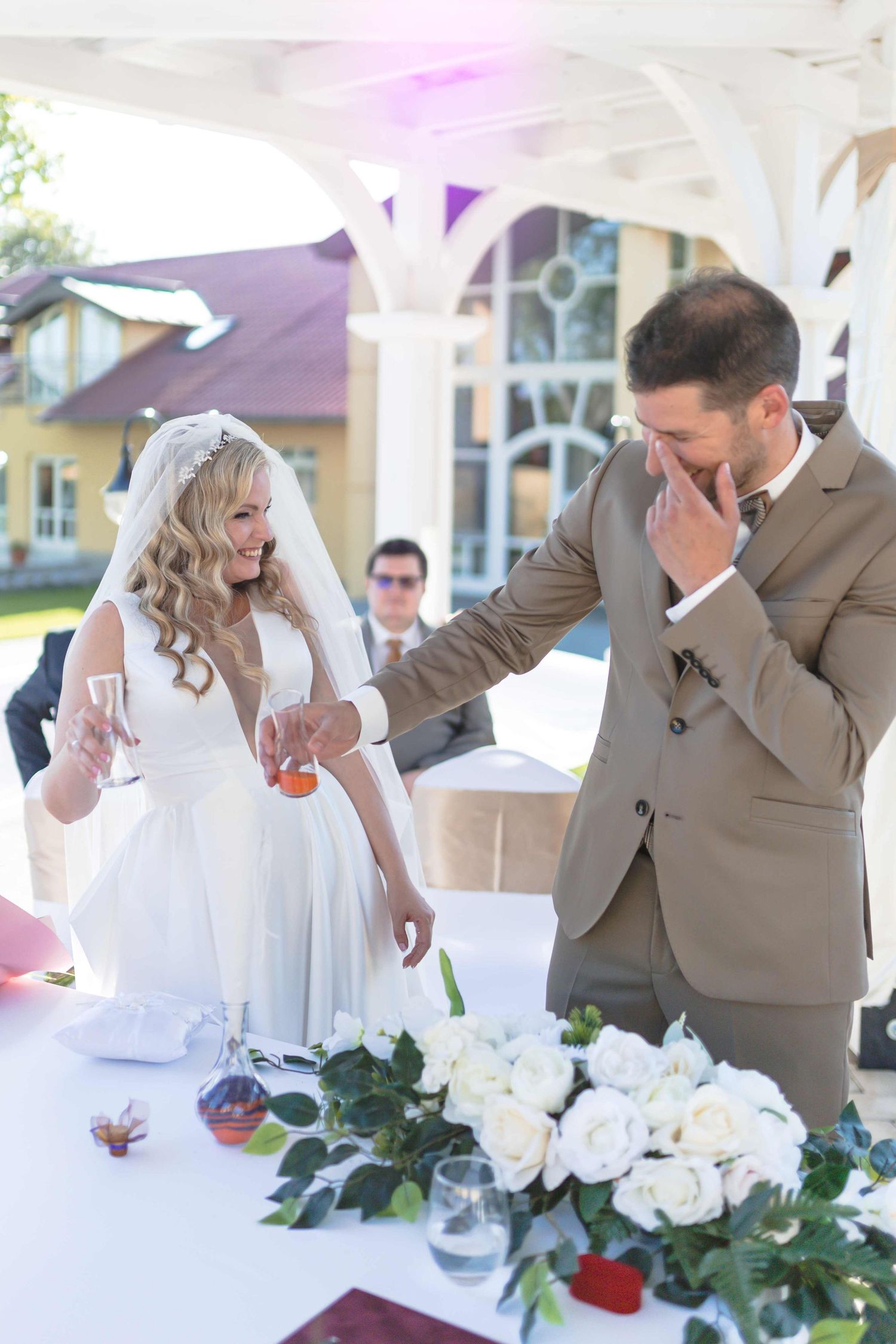 A joyful wedding moment captured at an outdoor reception with white roses and decorative floral arrangements on the table.