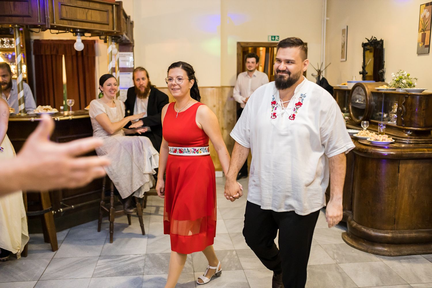 A couple enters a restaurant dining room while someone takes a photo, with the woman wearing a red dress.