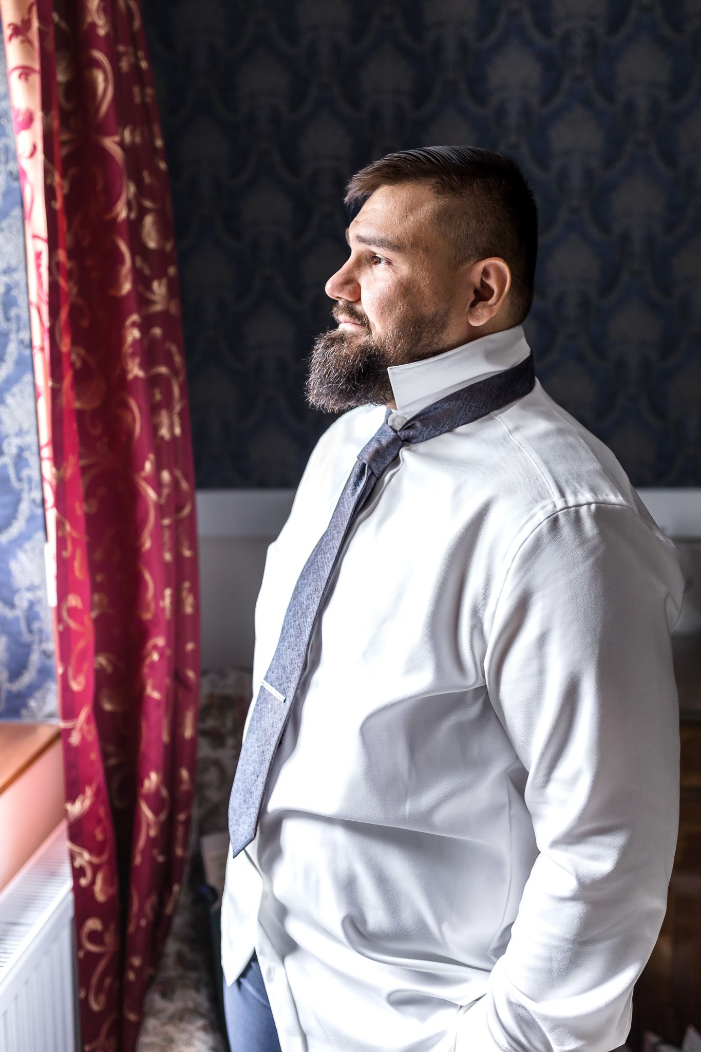 Side profile views of a person in a crisp white dress shirt against a dark patterned wall with red curtains.