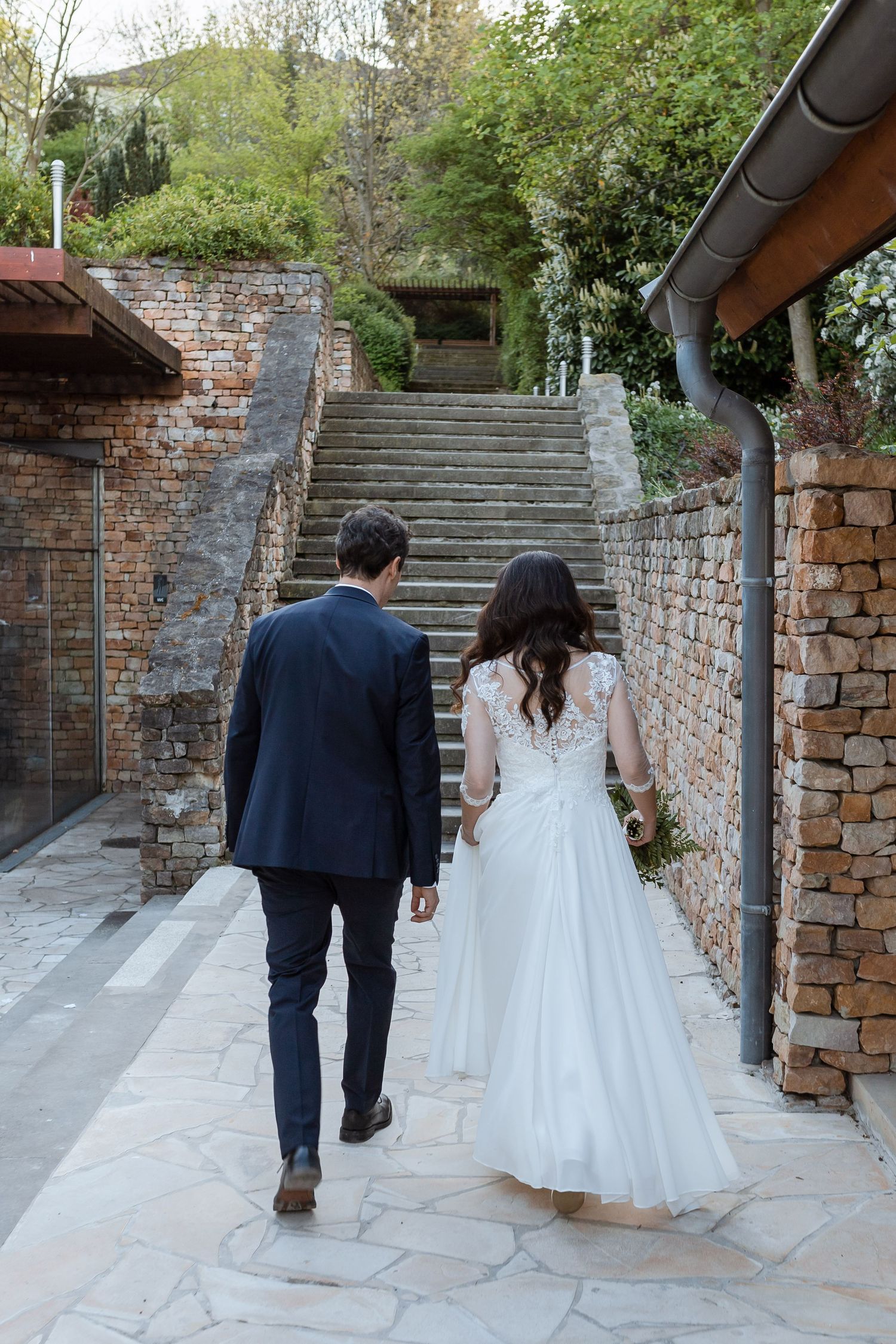 A couple in wedding attire walking up stone steps along a rustic brick wall in a garden setting.