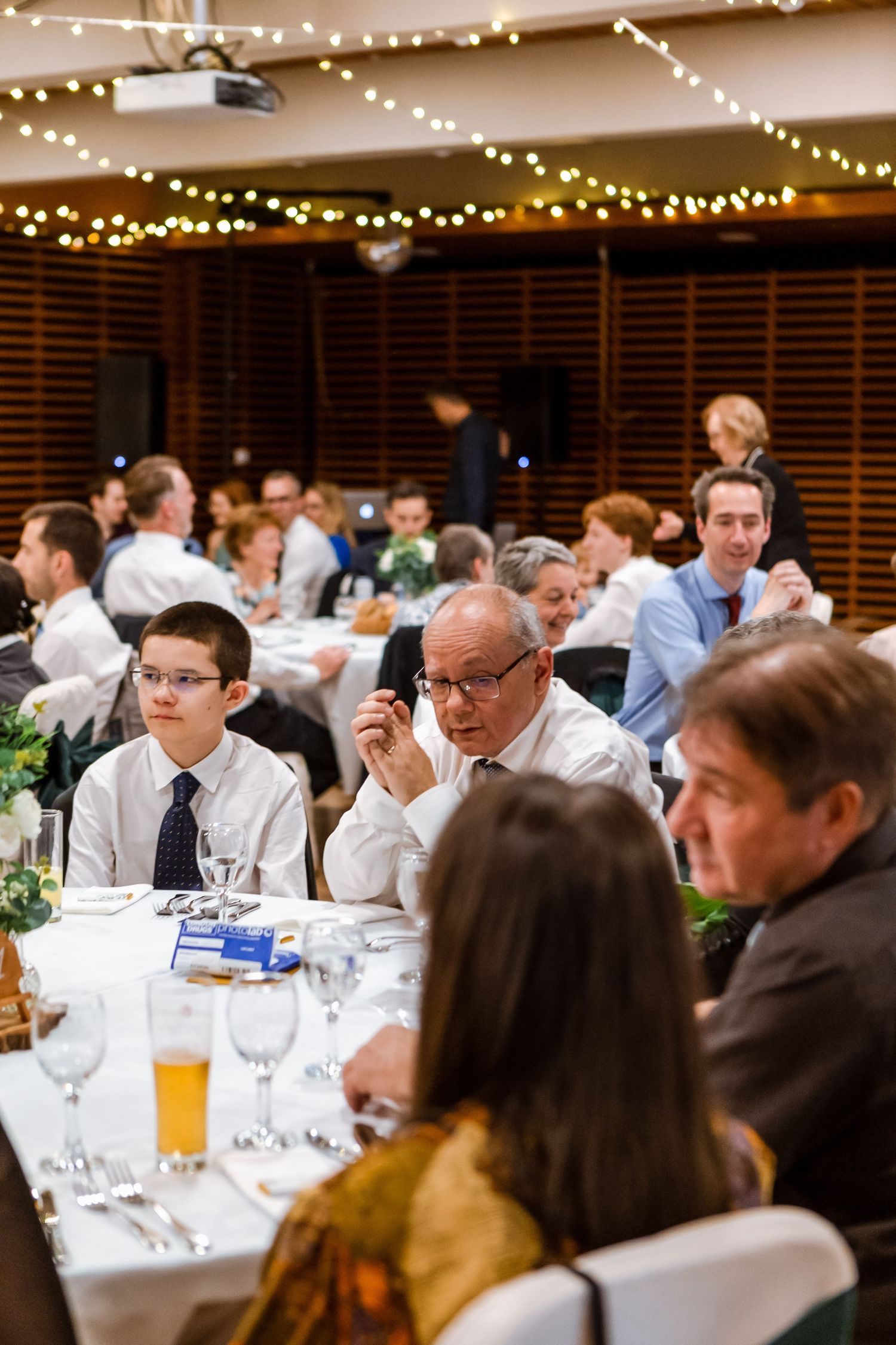 Guests seated at elegant round tables with white linens during a formal dinner event under warm string lights.