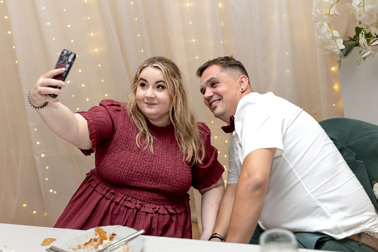 A couple takes selfies and poses together in front of a glowing string light backdrop at a festive event.