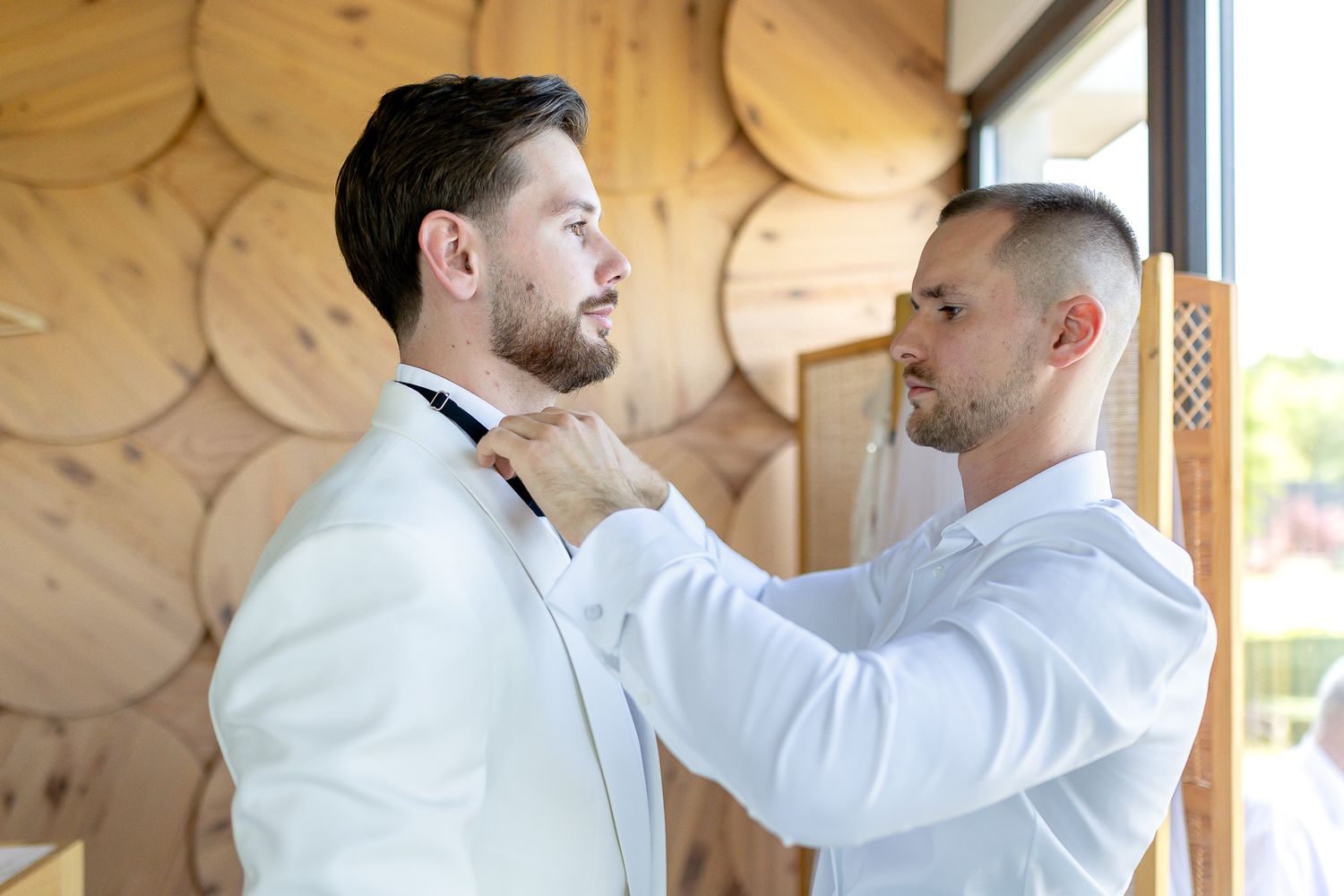 Two people in white formal wear embrace in front of a decorative wooden wall.