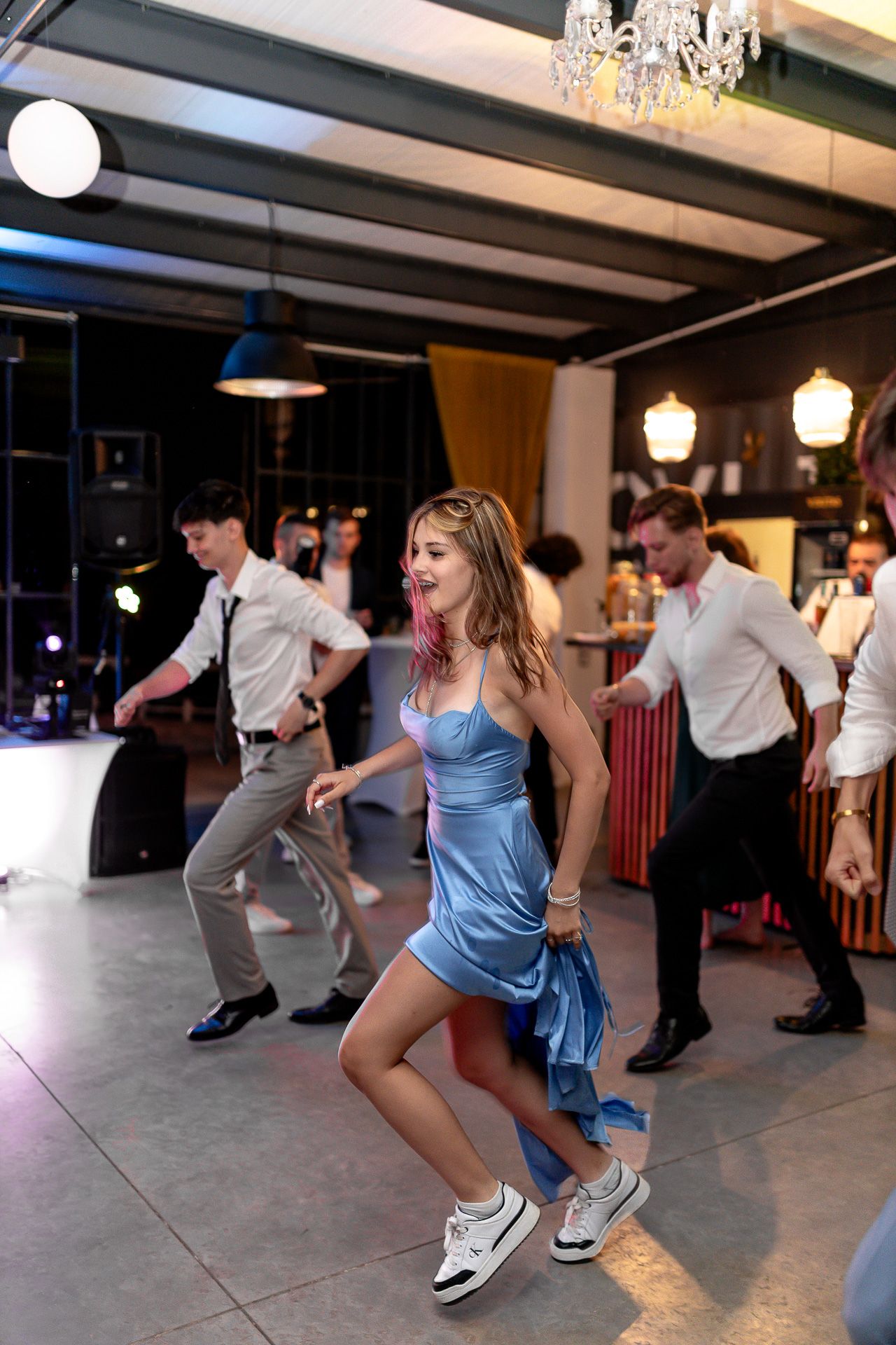 People dancing in casual attire under a chandelier at an indoor venue.