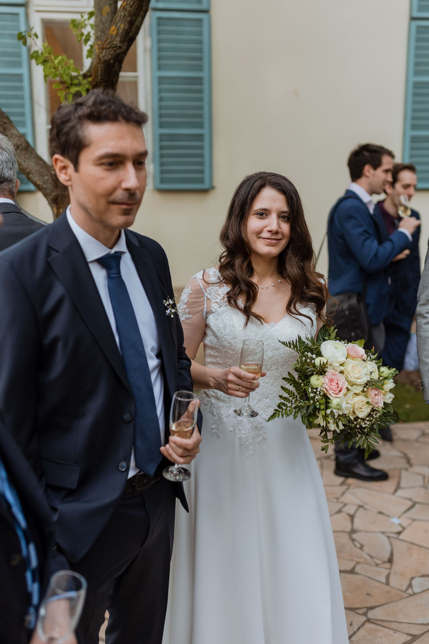 Newlywed couple shares intimate moments in garden setting with white dress and navy suit during outdoor reception.