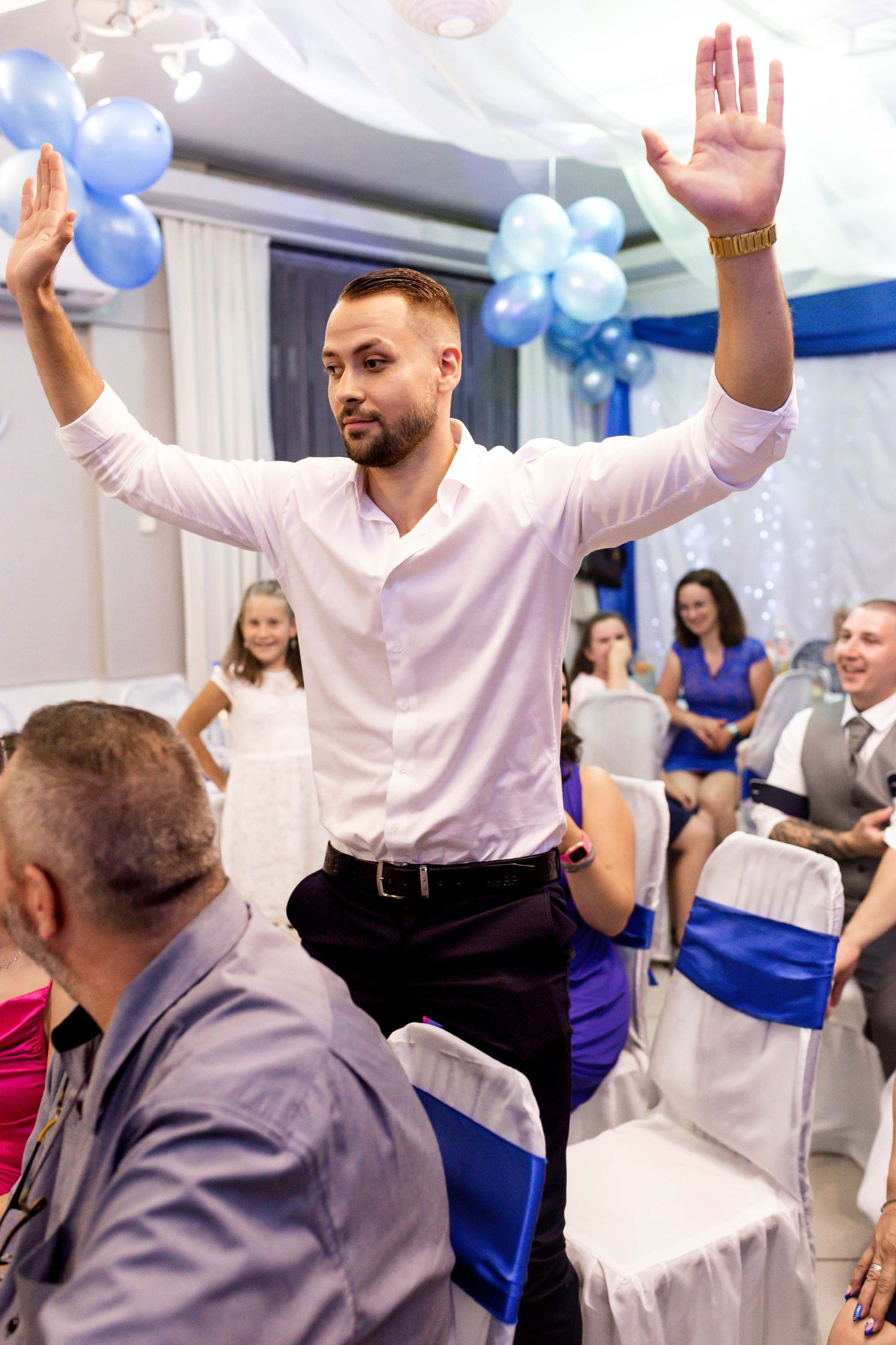 A person stands with raised arms at a celebration decorated with blue balloons and white chair covers in an indoor venue.