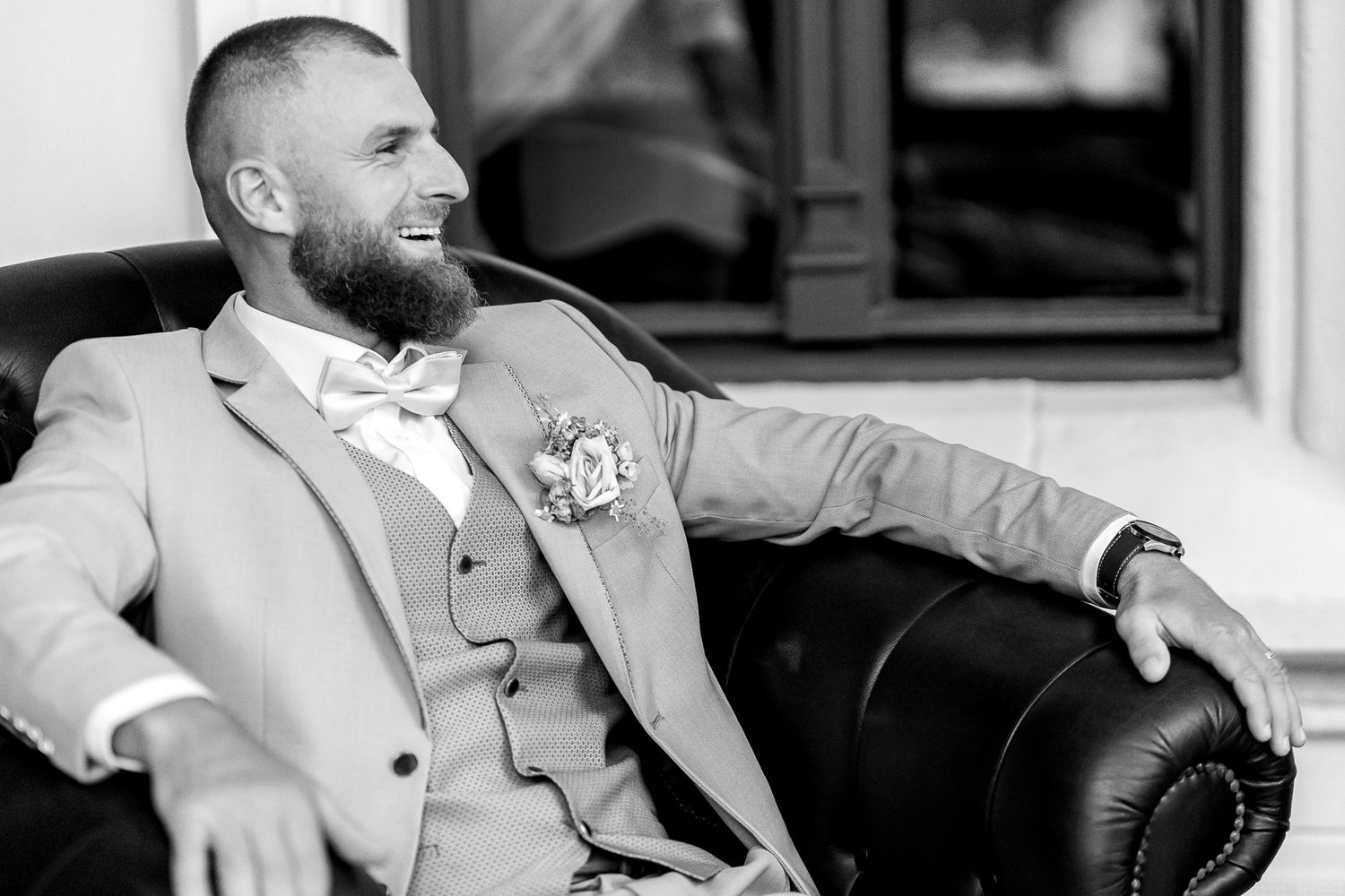 A groom in a light colored suit and boutonniere relaxes on a leather chair in black and white photography.