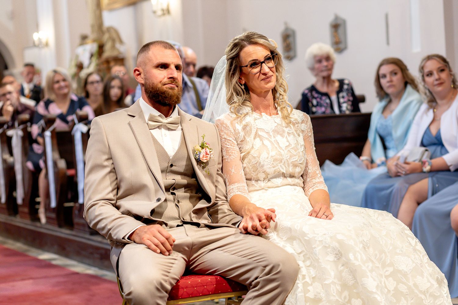 A wedding couple sits on wooden church pews during their ceremony surrounded by guests and natural light.