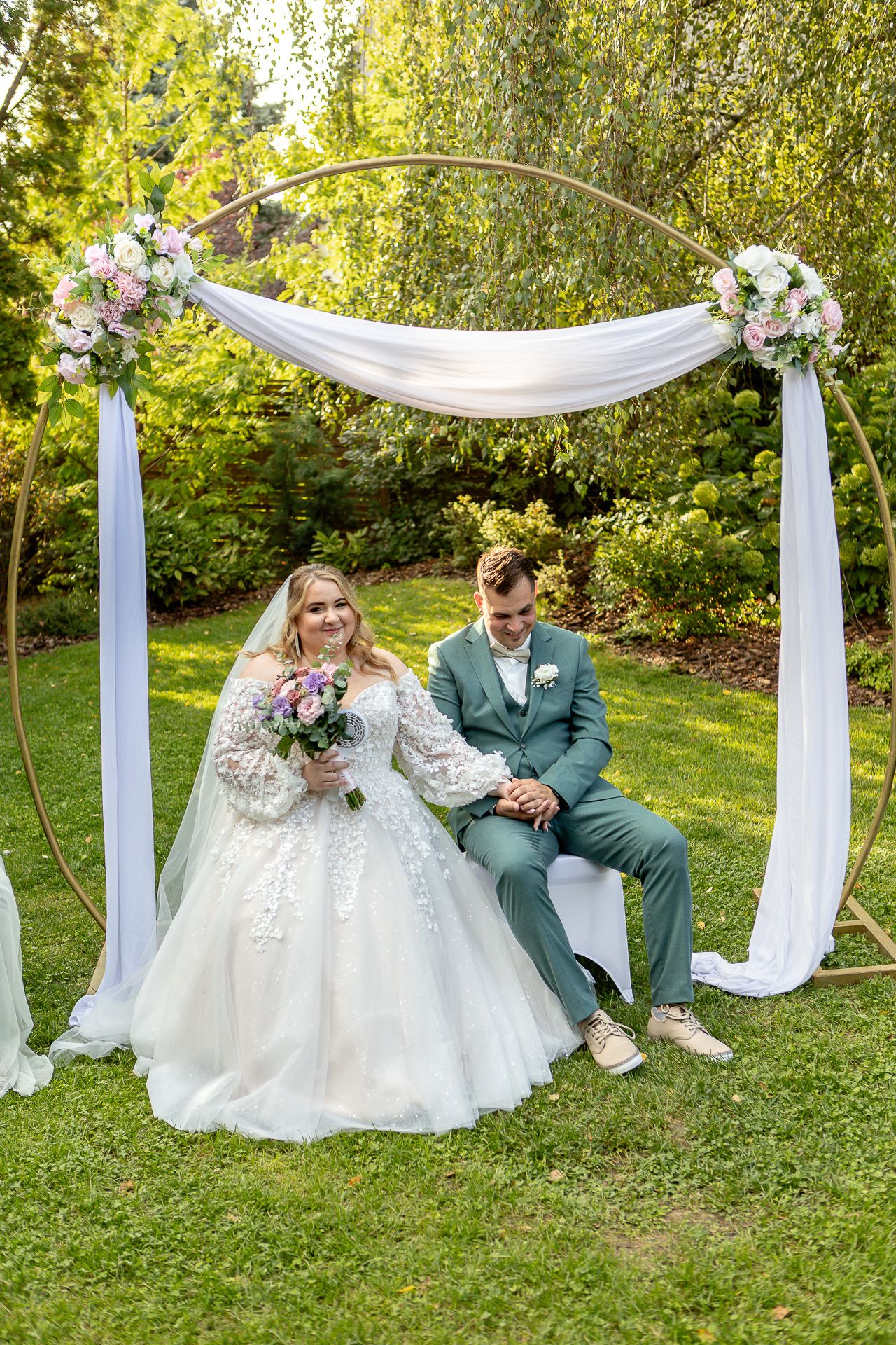 Couple sits together on grass under elegant white fabric draping between pillars in an outdoor garden setting.