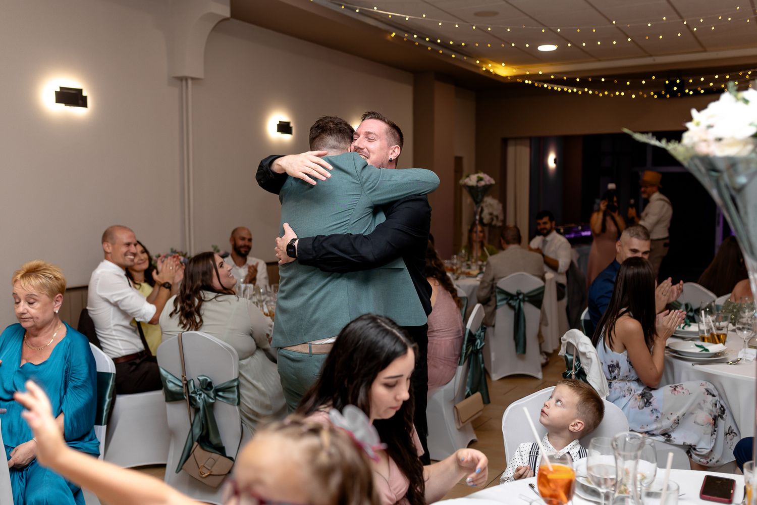 Two people embrace in a warm hug at a wedding reception while other guests sit at decorated tables with string lights above.