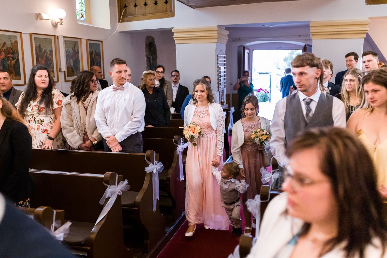 Wedding guests gather in wooden church pews decorated with white bows during a ceremony.