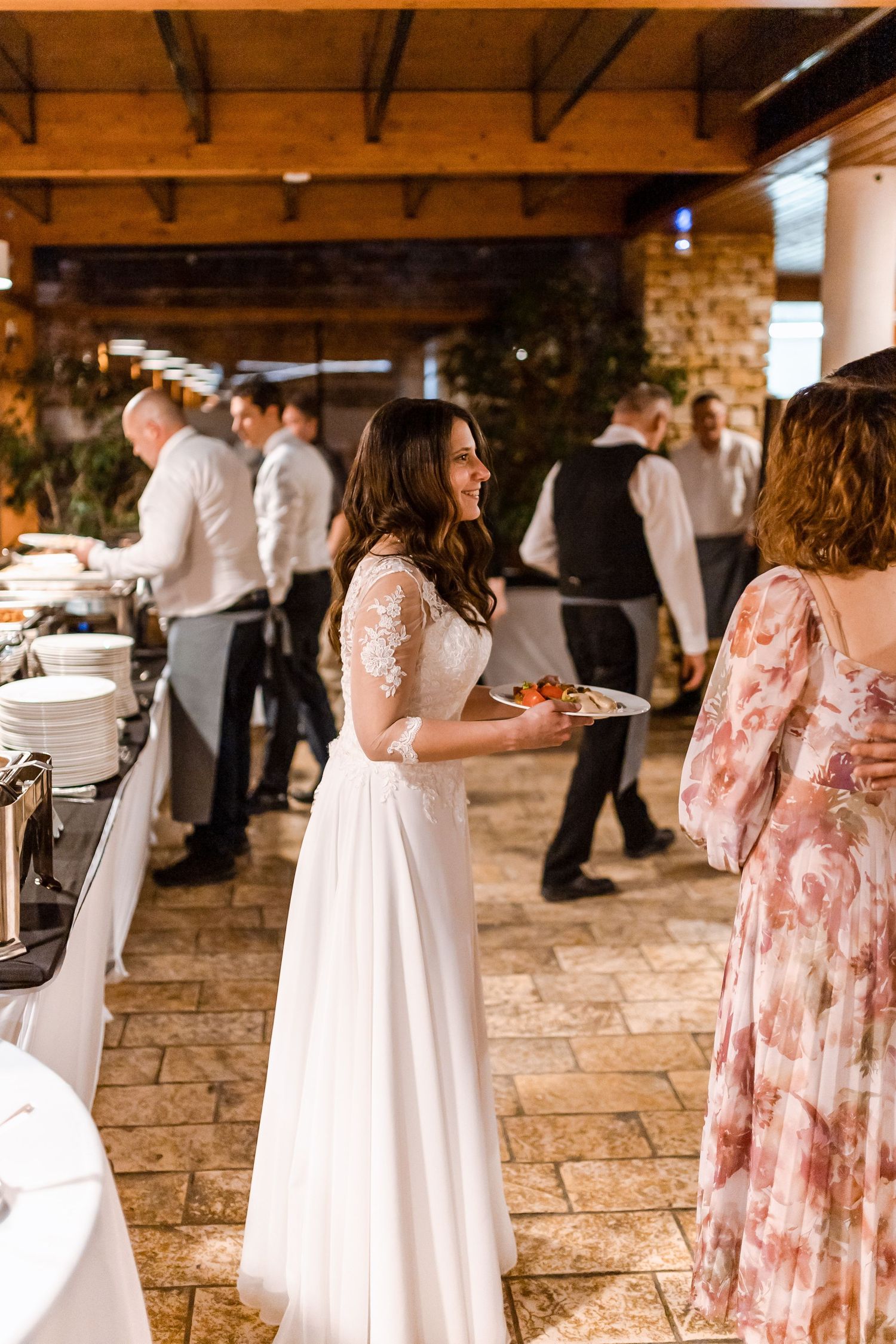 Wedding guests in white and floral pink dresses mingle at an elegant reception with stone floors and wooden beams.