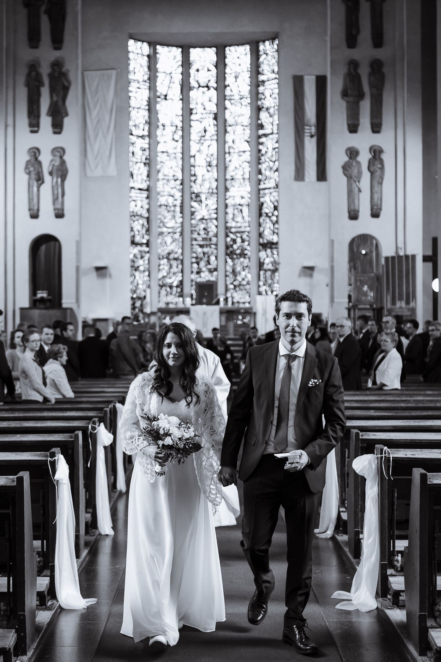 A bride and groom walk down the church aisle together after their ceremony, with stained glass windows visible behind them.