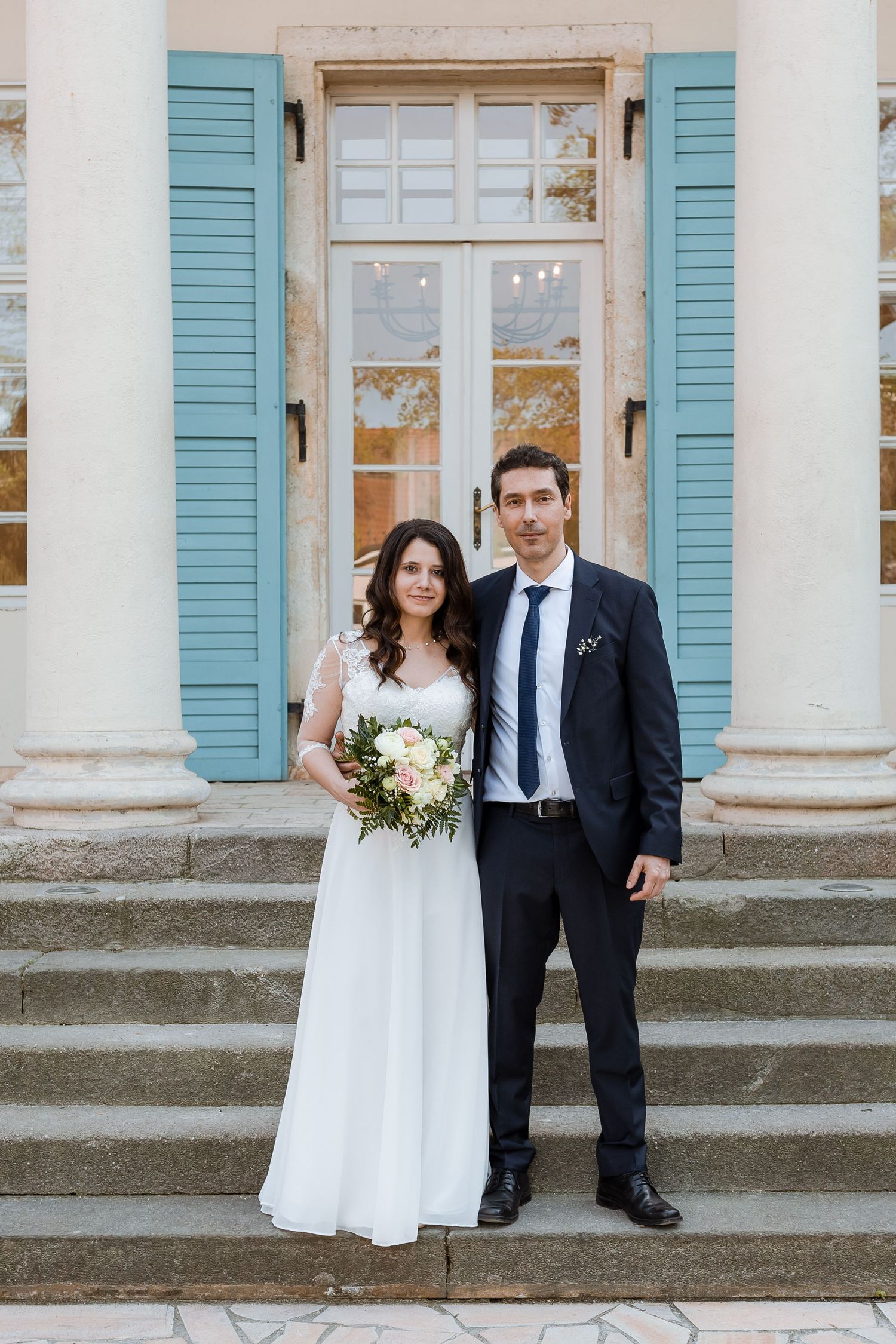 Wedding couple poses on historic mansion steps with white columns and blue shutters in elegant outdoor portrait.