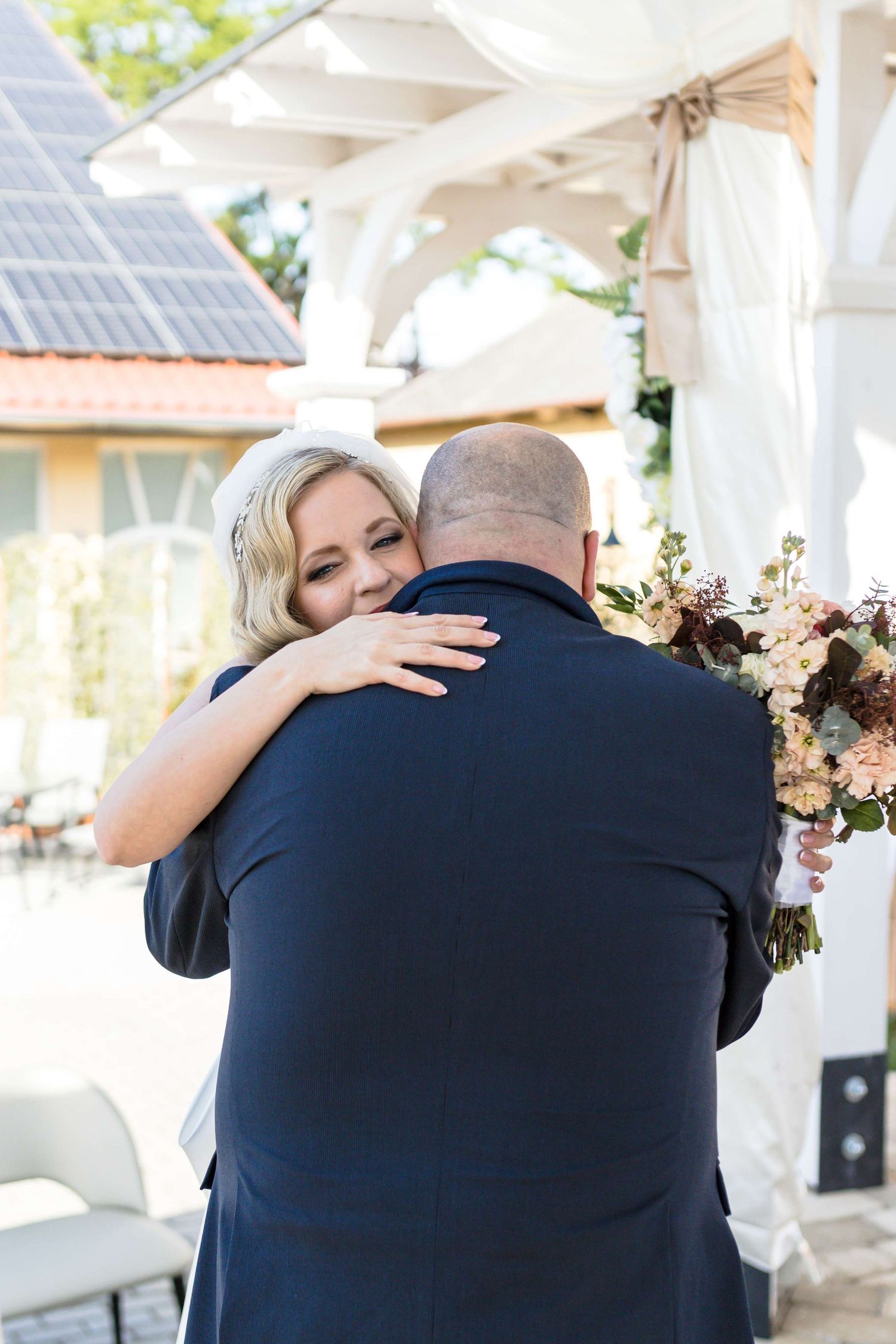 A tender emotional moment captured at an outdoor wedding venue with pink floral arrangements and white gazebo.