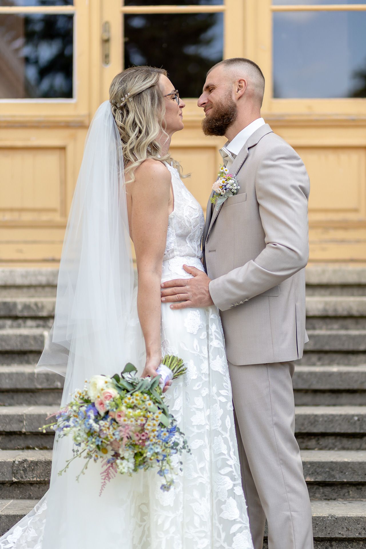 A romantic wedding couple embraces on stone steps, with the bride holding a pastel bouquet and wearing a flowing white gown.