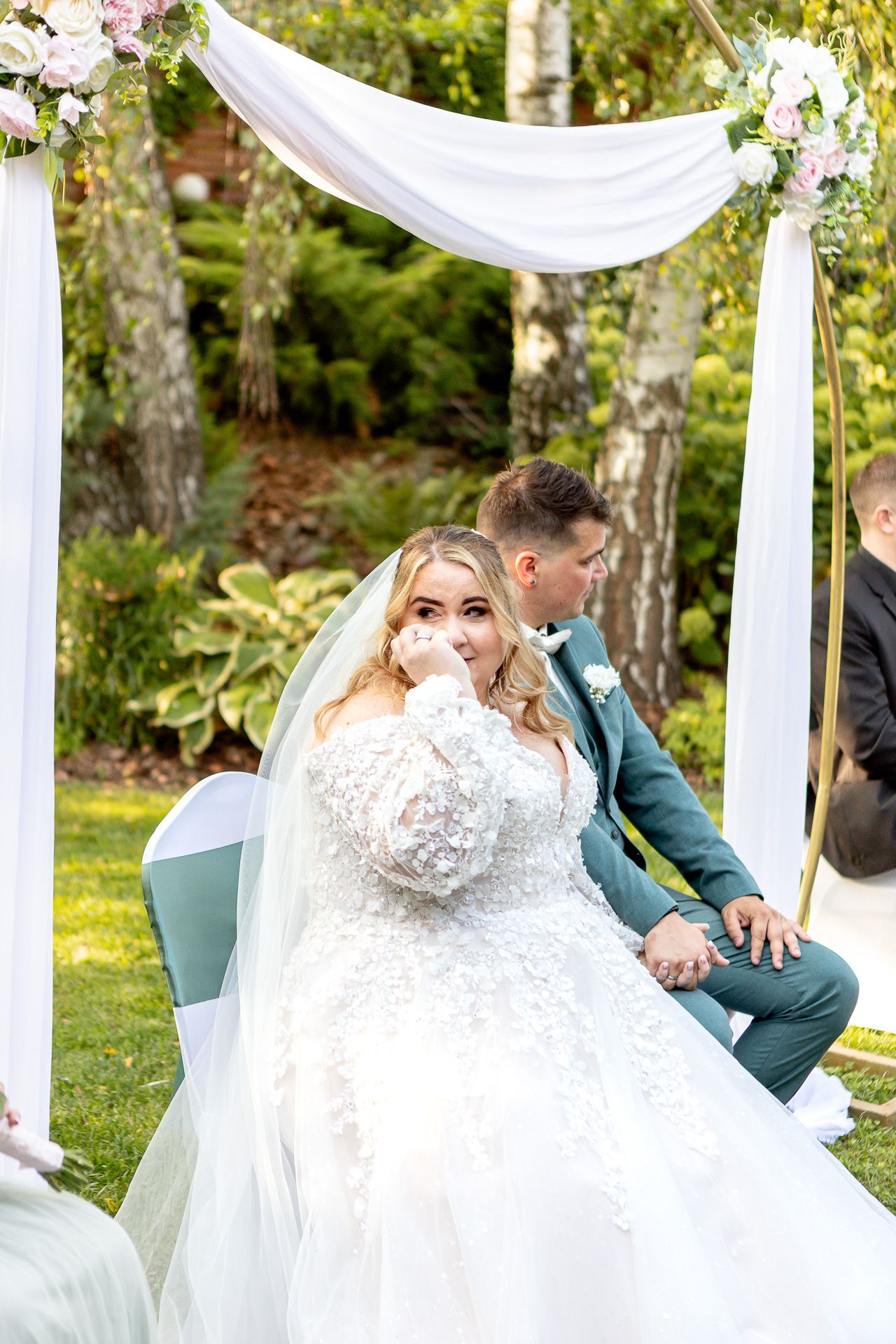 A couple shares a playful moment under white fabric draping at an outdoor garden wedding ceremony.
