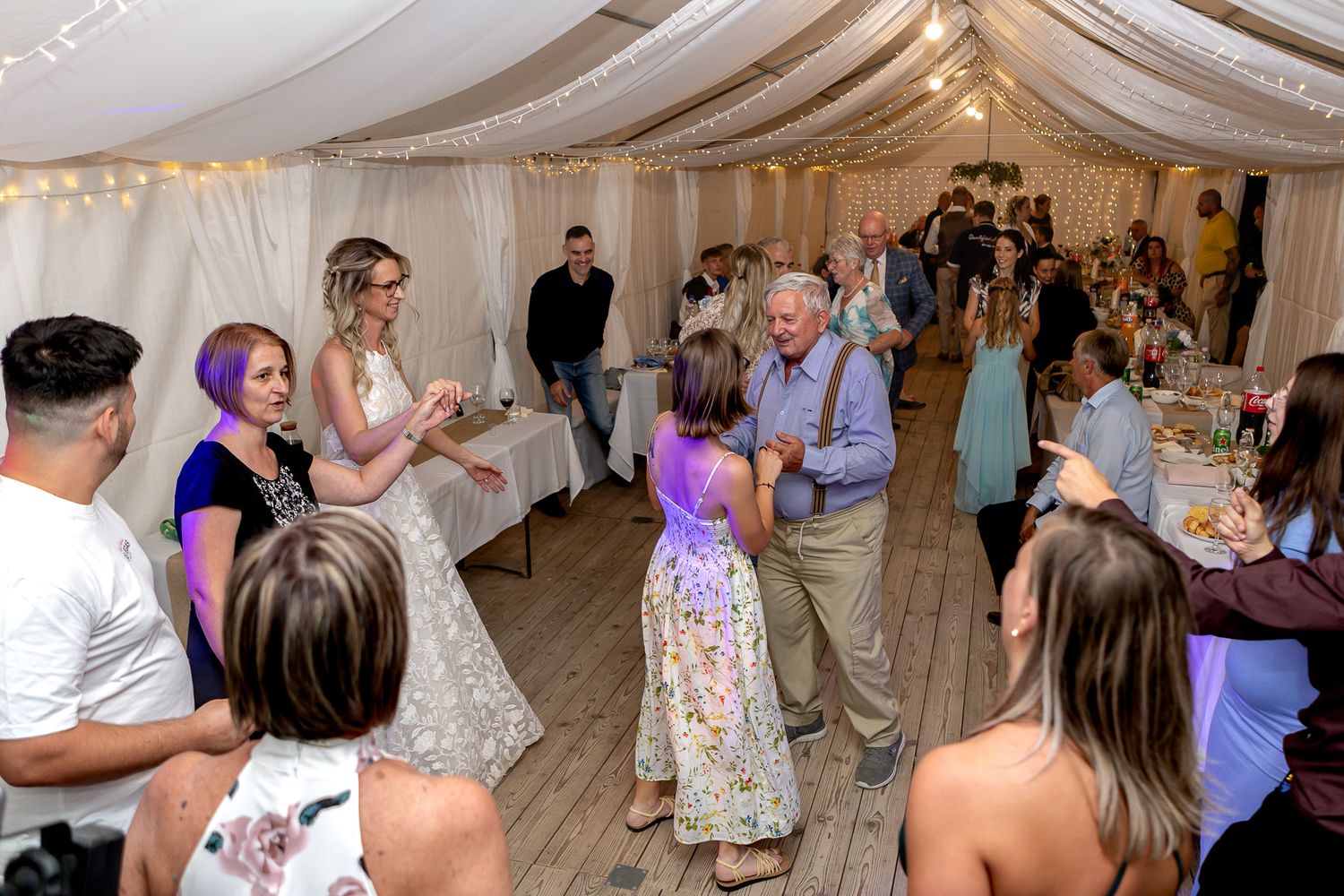 Wedding guests dance and celebrate together on a white-draped reception dance floor under string lights.