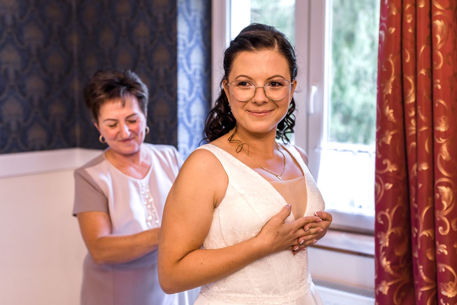 A heartfelt embrace takes place in a room with ornate wallpaper and red drapery during a special occasion.