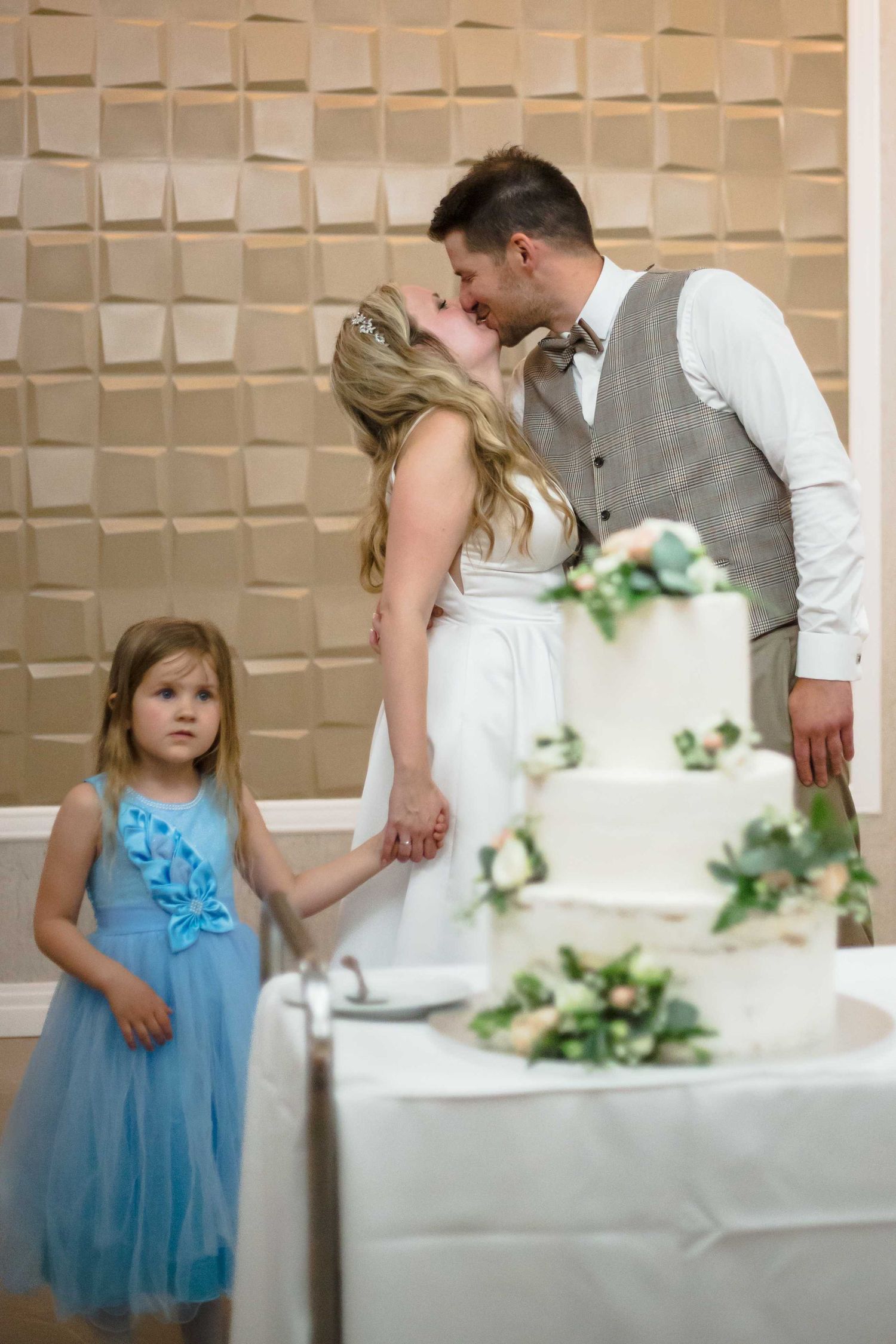 Wedding couple shares a sweet moment cutting their white tiered cake while a young attendant in blue looks on.