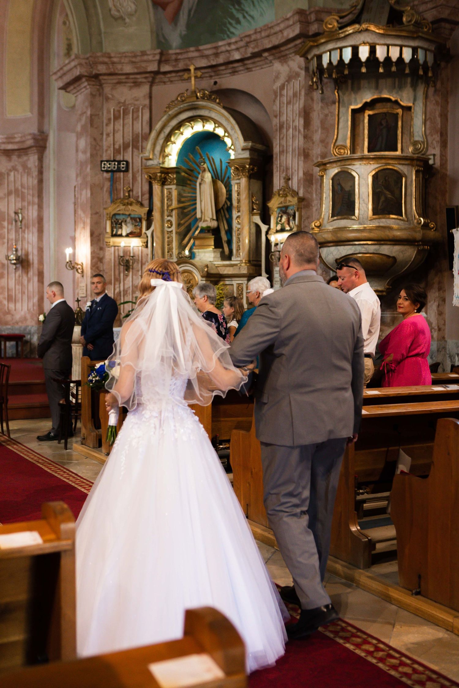 A wedding ceremony unfolds in a beautiful ornate church with white and gold architectural details and red carpeting.