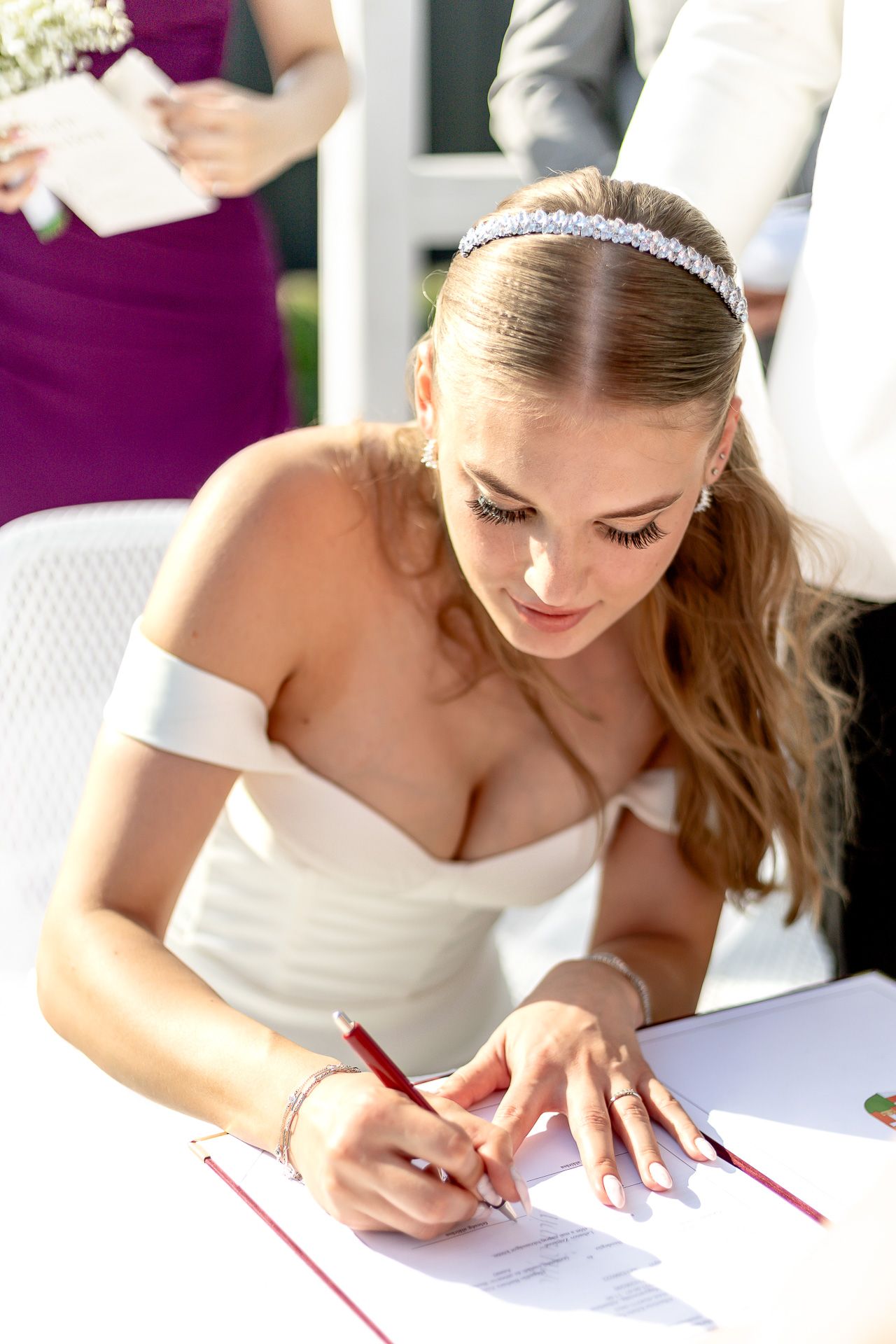 Bride in white dress signs marriage documents during outdoor ceremony.