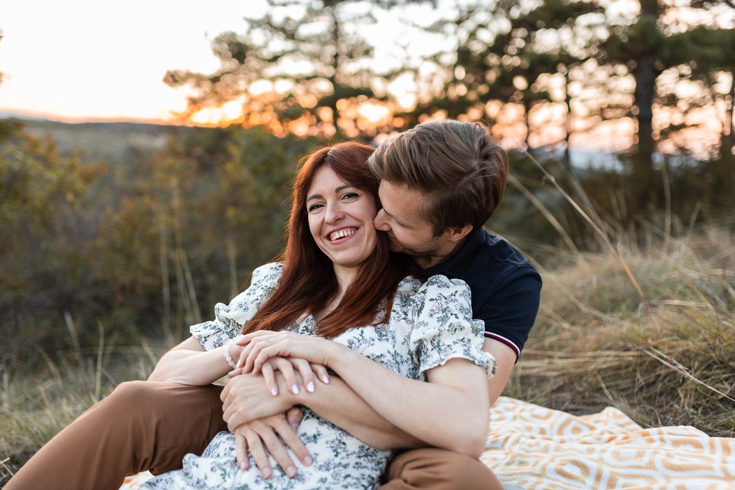 Romantic couple embracing on grassy hillside during sunset with trees in background.