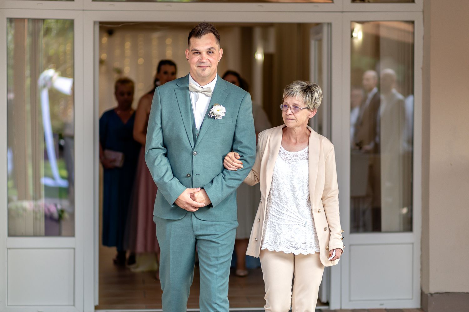A couple walks arm in arm through a doorway, with the person wearing a mint green suit and white name tag.