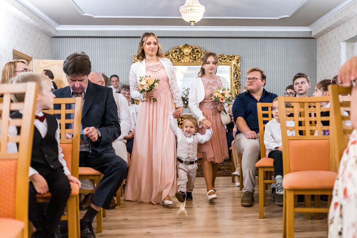 Wedding guests seated at wooden tables in a reception hall with peach-colored decor and ornate chandeliers.