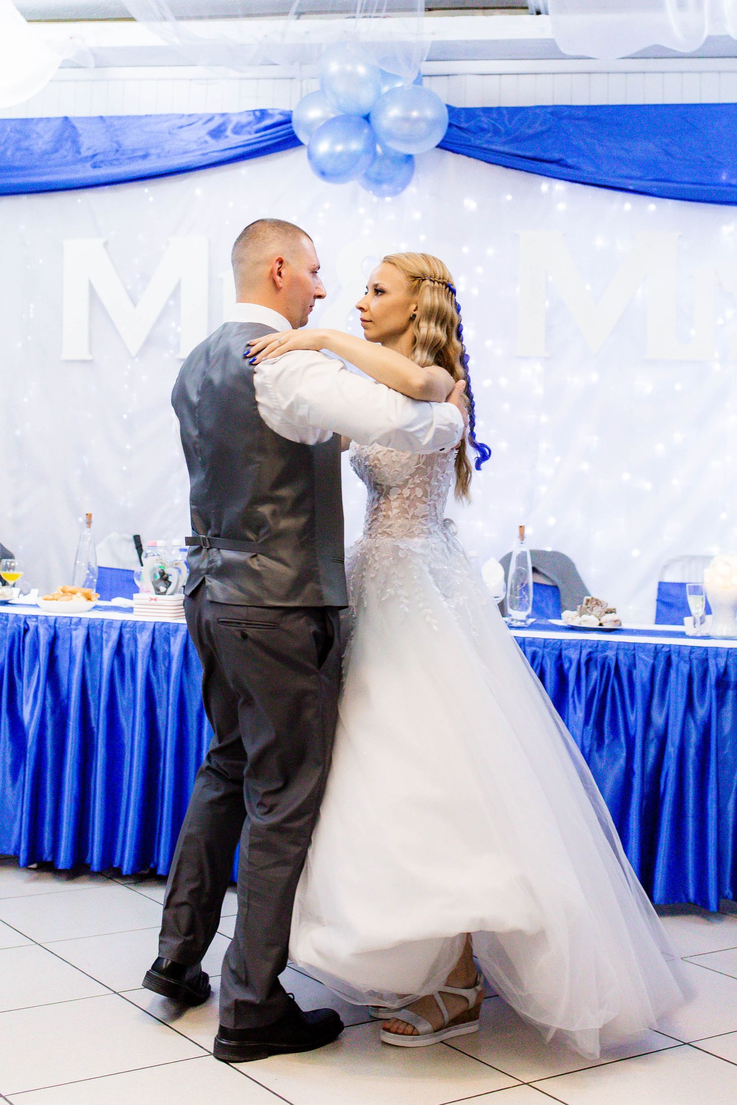 A couple shares their first dance at their wedding reception with blue and white decor and a decorated head table.