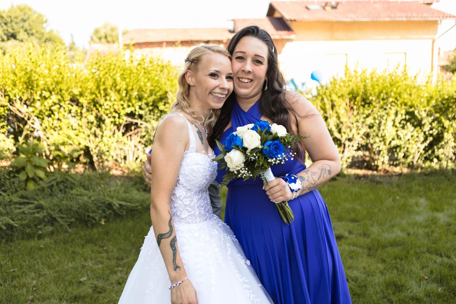 Two people in wedding attire share a joyful moment in a garden setting with bright sunlight and greenery.