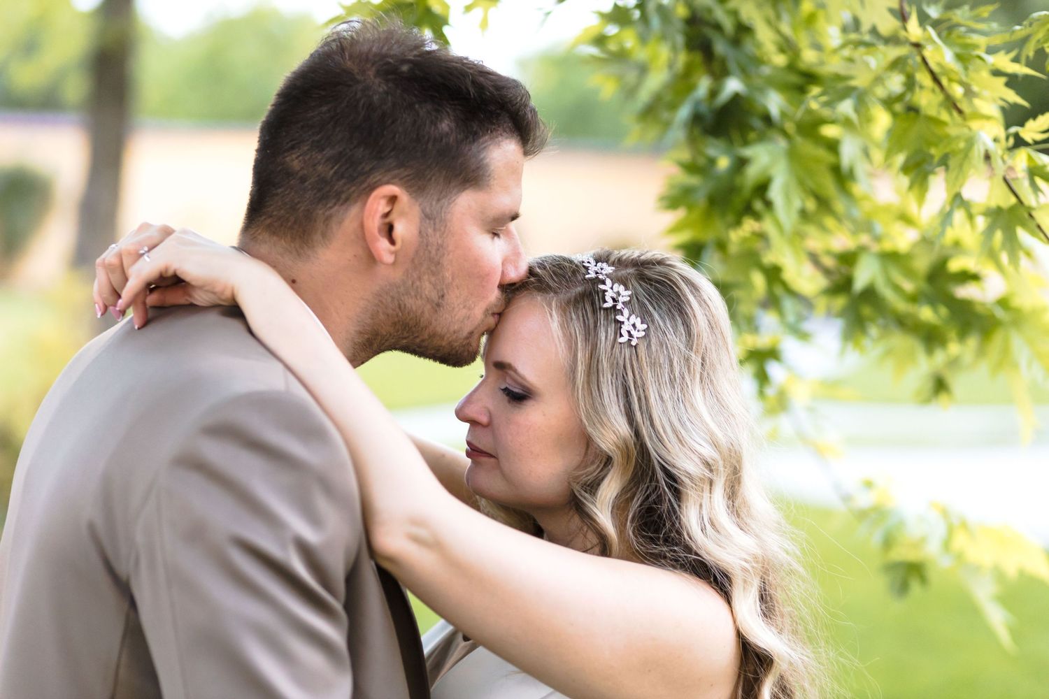 A romantic couple shares tender kisses in a sunlit outdoor setting with green foliage in the background.